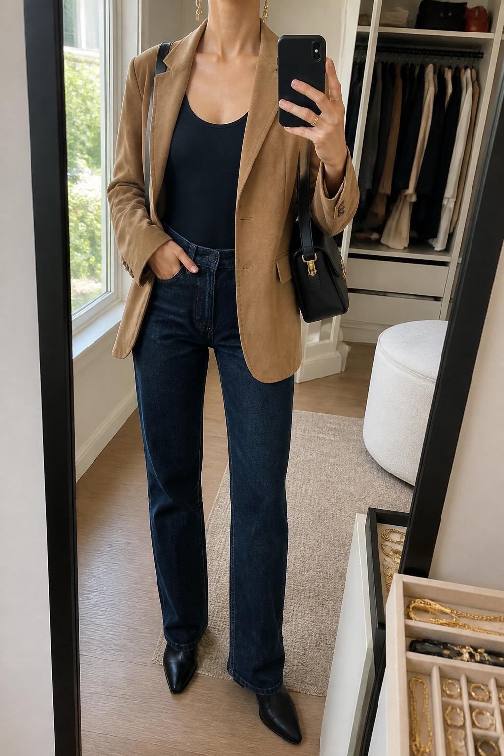 mirror selfie of a woman wearing the exact outfit described above, clearly showing a camel suede blazer layered over a black fitted bodysuit, dark blue straight-leg jeans, black pointed ankle boots, a black leather shoulder bag, and gold drop earrings, holding a phone in front of a mirror, cropped at NECK so the face does not show, with a relaxed casual selfie pose, in a walk-in closet mirror, natural window light, with an open closet with clothes visible and a jewelry tray on the vanity, latest iPhone photo quality.