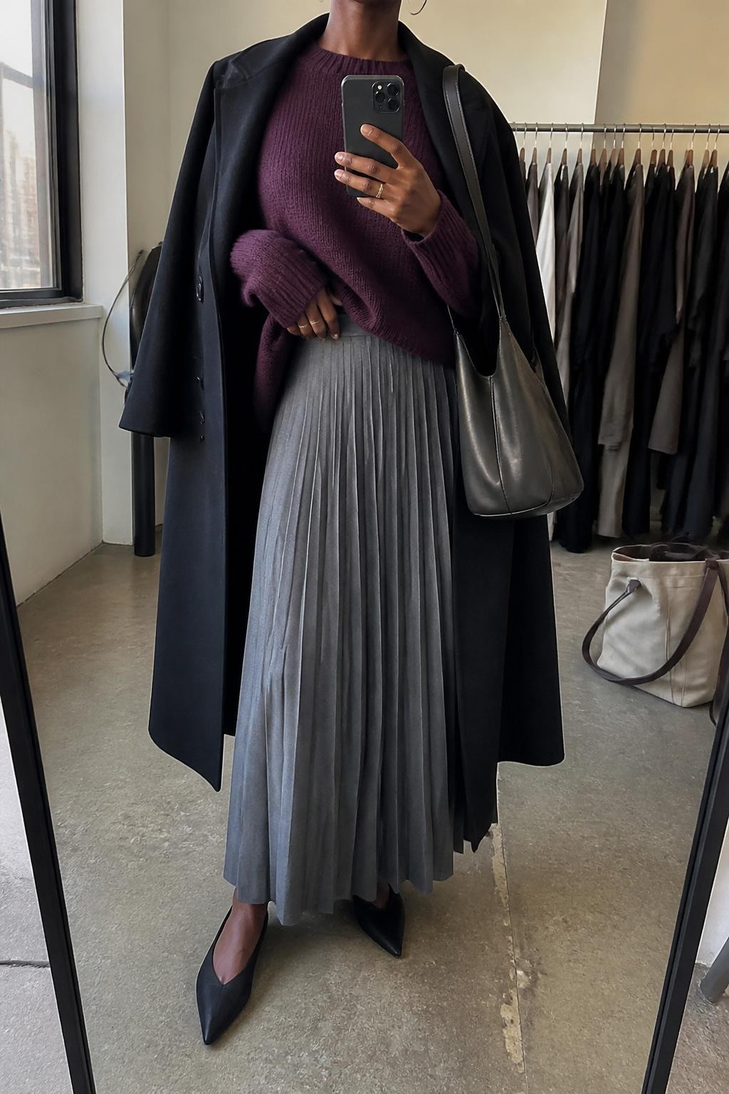 mirror selfie of a Black woman wearing the exact outfit described above, clearly showing a gray pleated maxi skirt, plum oversized knit sweater tucked slightly at the front, black pointed flats, charcoal shoulder bag, silver hoop earrings, and black wool coat draped open, holding a phone in front of a mirror, cropped at NECK so the face does not show, with a relaxed casual selfie pose, in a fashion studio mirror corner, natural window light, with clothing rack with garments behind and a tote bag resting on the floor, latest iPhone photo quality.