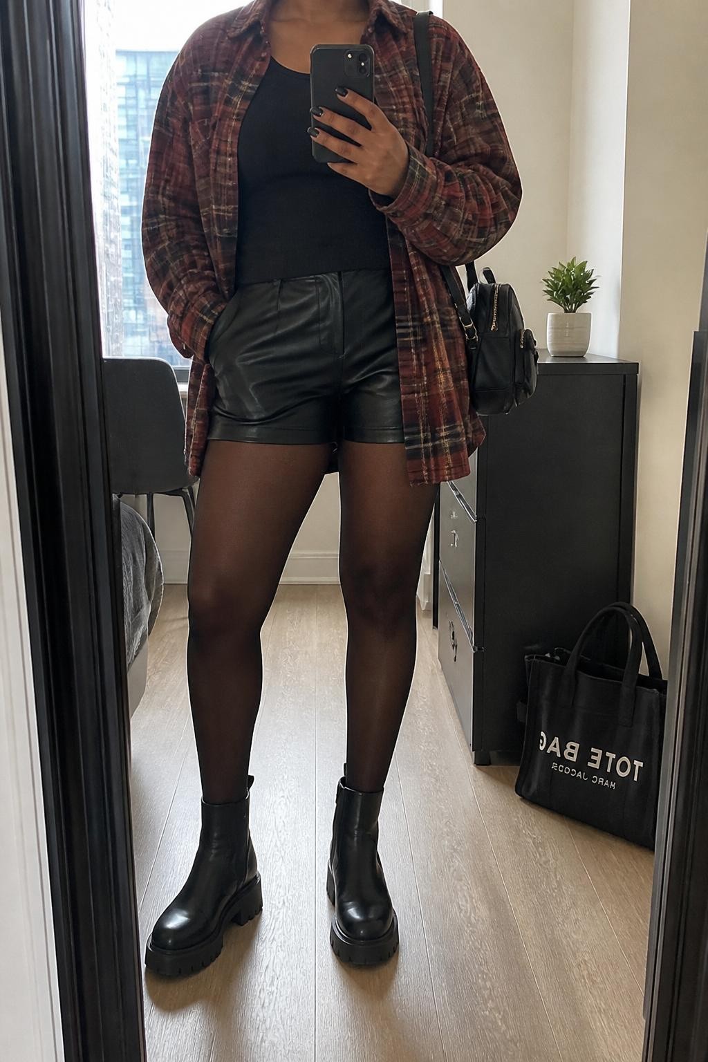 mirror selfie of a Black woman wearing the exact outfit described above, clearly showing an oversized red and brown plaid shirt worn open, fitted black ribbed tank, black leather shorts, sheer black tights, black chunky ankle boots, and a black mini backpack purse, holding a phone in front of a mirror, cropped at NECK so the face does not show, with a relaxed casual selfie pose, in a stylish city apartment, natural window light, with a small plant on a dresser and a tote bag resting on the floor, latest iPhone photo quality.
