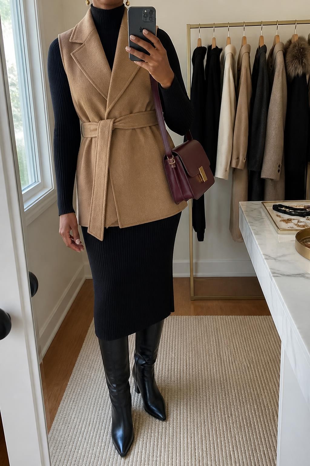 Mirror selfie of a Black woman wearing a camel wool-blend belted vest over a fitted black ribbed-knit midi dress, black leather knee-high boots, a structured burgundy shoulder bag, and small gold drop earrings, clearly showing the exact clothing items, colors, fabrics, and accessories listed, holding a phone in front of a minimal dressing room mirror, cropped at the neck so the face does not show, relaxed confident selfie pose, natural window light, a clothing rack with garments behind and a jewelry tray on the vanity, polished and uncluttered, latest iPhone photo quality.