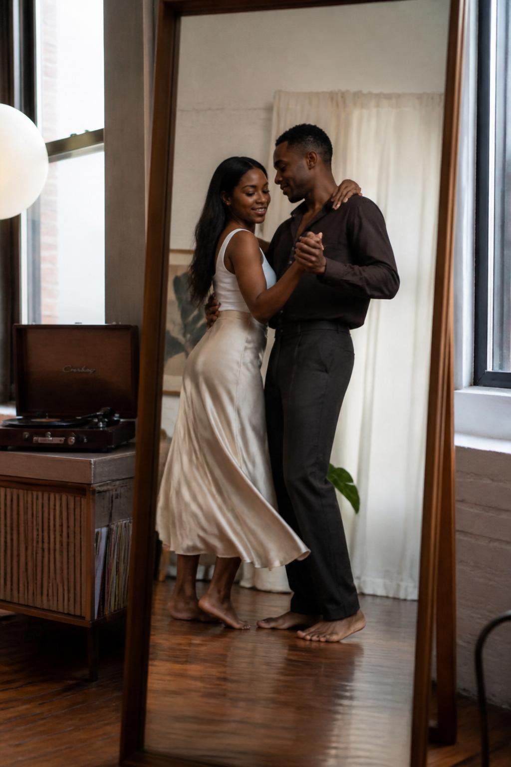 Image prompt: A romantic indoor couple photo reflected in a tall wooden mirror in a loft apartment, featuring an adult Black woman with deep brown skin wearing a soft champagne satin skirt and fitted white tank, dancing slowly with her partner in dark trousers and an open-collar shirt, her hand placed on his shoulder, his hand holding hers gently at chest height, both reflected in the mirror with their bodies slightly turned, bare feet on warm wooden floors, record player on a cabinet, soft curtain-filtered daylight, a half-open window, natural motion blur in the skirt, realistic skin texture, gentle smiles, modern romantic lifestyle photography, human and polished.