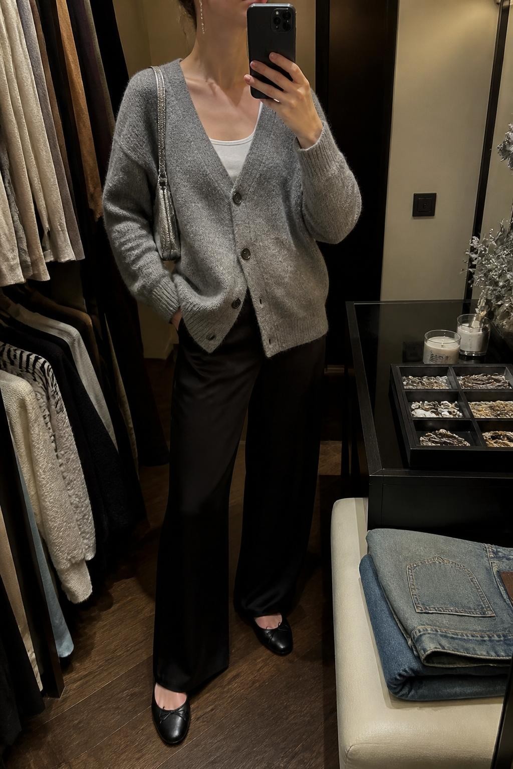 Mirror selfie of a woman wearing a soft gray oversized cardigan buttoned over a fitted white tank top and black satin wide-leg trousers, clearly showing black leather ballet flats, a small silver shoulder bag, and delicate silver drop earrings, holding an iPhone in front of a walk-in closet mirror, cropped at the neck so the face does not show, relaxed casual selfie pose, warm moody lighting, a jewelry tray on the vanity and folded denim jeans on a bench, natural unstaged setting, latest iPhone photo quality.