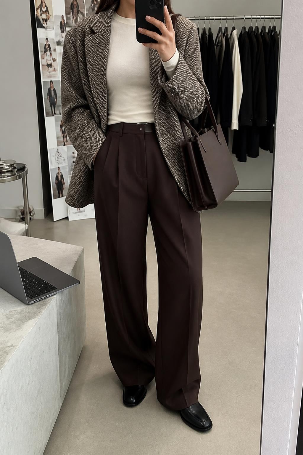 Mirror selfie of a woman wearing chocolate-brown tailored wide-leg trousers, an ivory fitted long-sleeve top, an oversized grey and brown herringbone wool blazer, black leather loafers, and a structured dark brown tote bag, clearly showing the exact clothing items, colors, fabrics, and accessories listed, holding a phone in front of a fashion studio mirror corner, cropped at the neck so the face does not show, relaxed casual selfie pose, soft indoor lighting, a laptop on a bench and a clothing rack with garments behind, natural professional setting, latest iPhone photo quality.