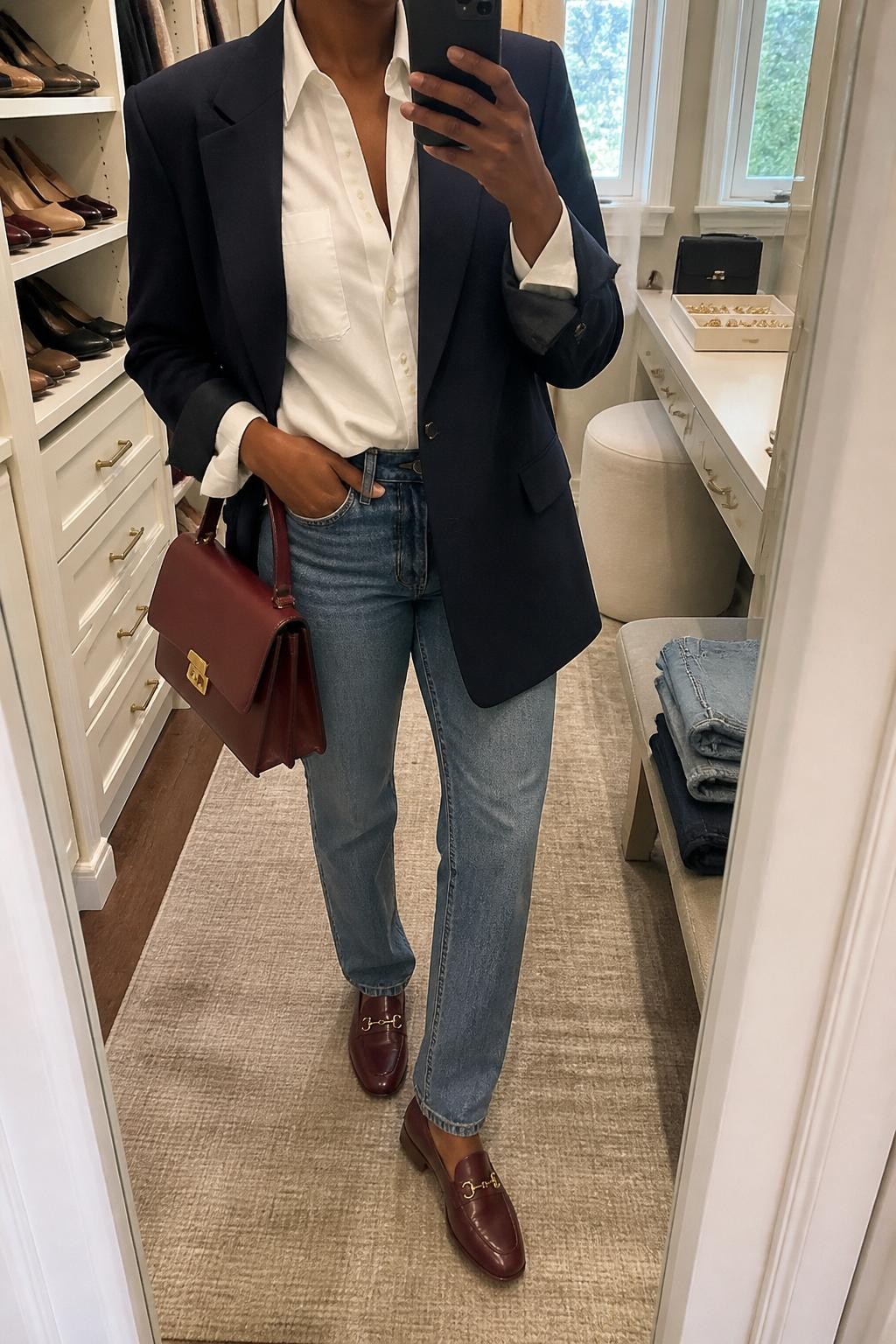 mirror selfie of a Black woman wearing the exact outfit described above, clearly showing a navy oversized blazer layered over a crisp white button-down shirt, medium-wash straight-leg jeans, burgundy loafers, a burgundy top-handle bag, and pearl stud earrings, holding a phone in front of a mirror, cropped at NECK so the face does not show, with a relaxed casual selfie pose, in a walk-in closet mirror, natural window light, with folded denim jeans on a bench and a jewelry tray on the vanity, latest iPhone photo quality.