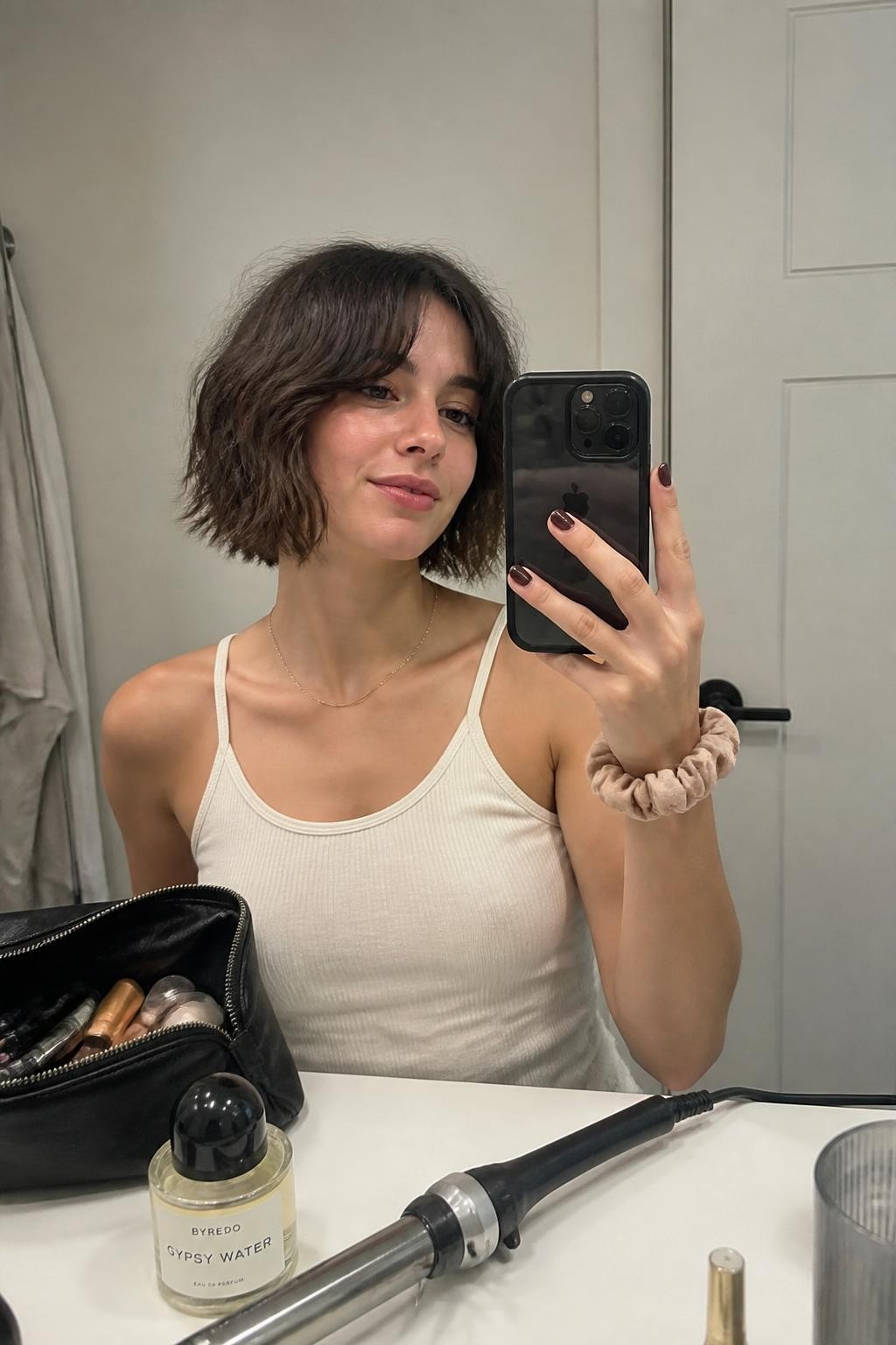 Image prompt: Photo of a woman wearing a tousled jaw-length shaggy bob with choppy layers and soft curtain bangs, candid mirror selfie, bathroom vanity mirror with a makeup bag and perfume bottle nearby, soft indoor lighting, containing a curling wand on the counter and a scrunchie around her wrist, realistic casual getting-ready moment, latest iPhone photo quality.