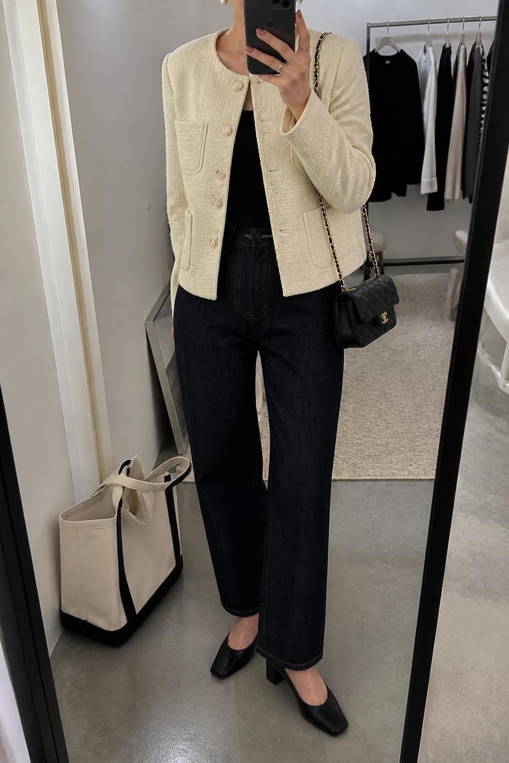 mirror selfie of a woman wearing the exact outfit described above, clearly showing a cream tweed jacket layered over a black shell top, dark denim trouser jeans, black block heel pumps, a black quilted mini bag, and pearl earrings, holding a phone in front of a mirror, cropped at NECK so the face does not show, with a relaxed casual selfie pose, in a fashion studio mirror corner, soft indoor lighting, with a tote bag resting on the floor and a clothing rack with garments behind, latest iPhone photo quality.