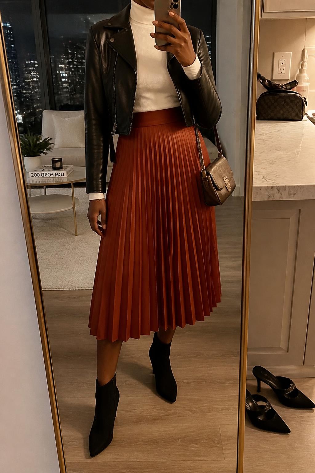 Mirror selfie of a Black woman wearing a rust-orange pleated midi skirt, fitted ivory turtleneck, cropped black leather jacket, black suede ankle boots, and a metallic bronze mini shoulder bag, clearly showing the exact clothing items, colors, fabrics, and accessories listed, holding a phone in front of a stylish city apartment mirror, cropped at the neck so the face does not show, relaxed casual selfie pose, warm moody lighting, a pair of heels on the floor nearby and a makeup bag on the counter, natural evening setting, latest iPhone photo quality.