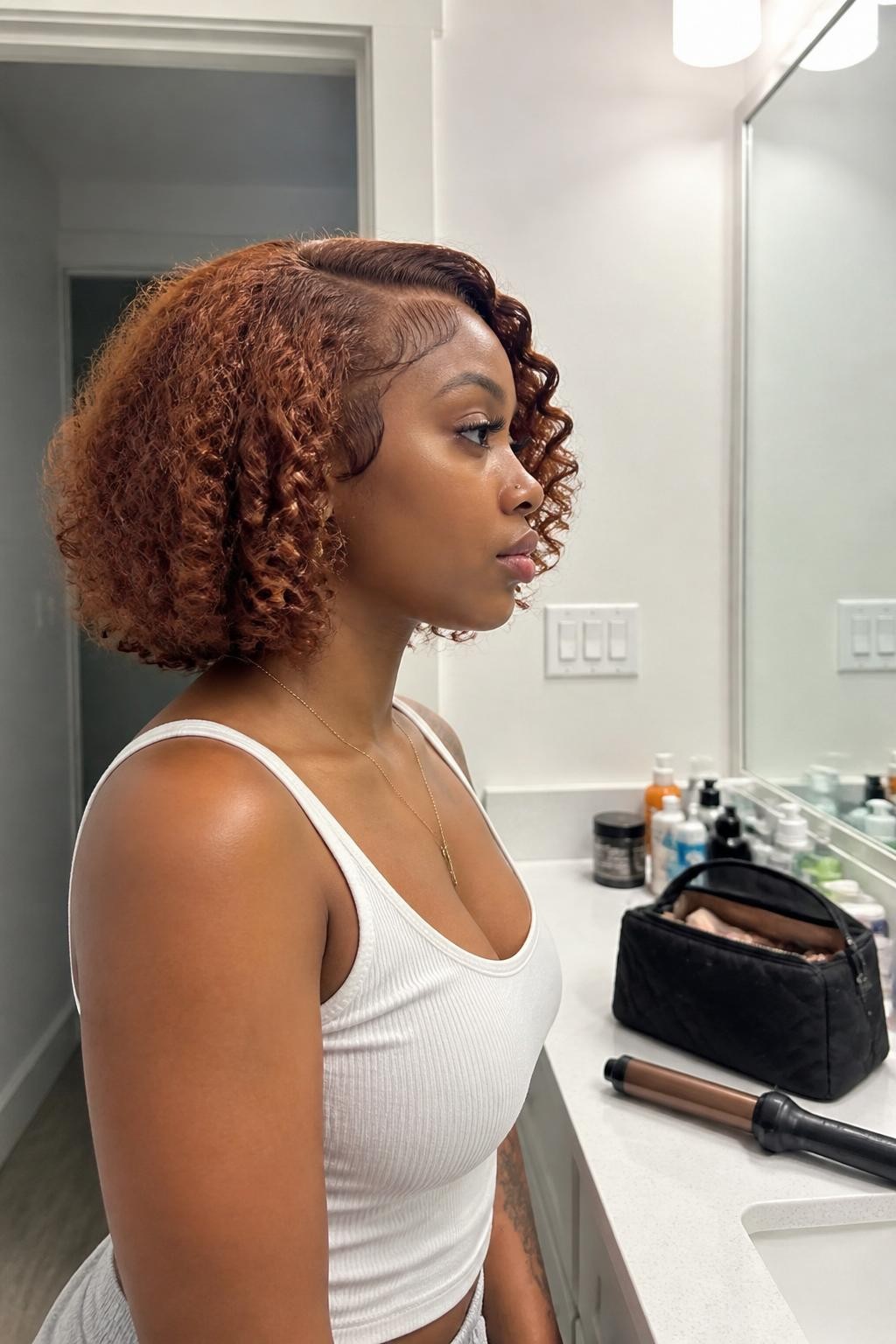 Image prompt: Photo of a Black woman wearing a rounded auburn crochet bob with soft defined curls and a gentle side part, side profile view, bathroom vanity mirror with skincare products and a makeup bag nearby, soft indoor lighting, containing small gold earrings peeking through and a curling iron resting unplugged on the counter, realistic getting-ready moment, latest iPhone photo quality.
