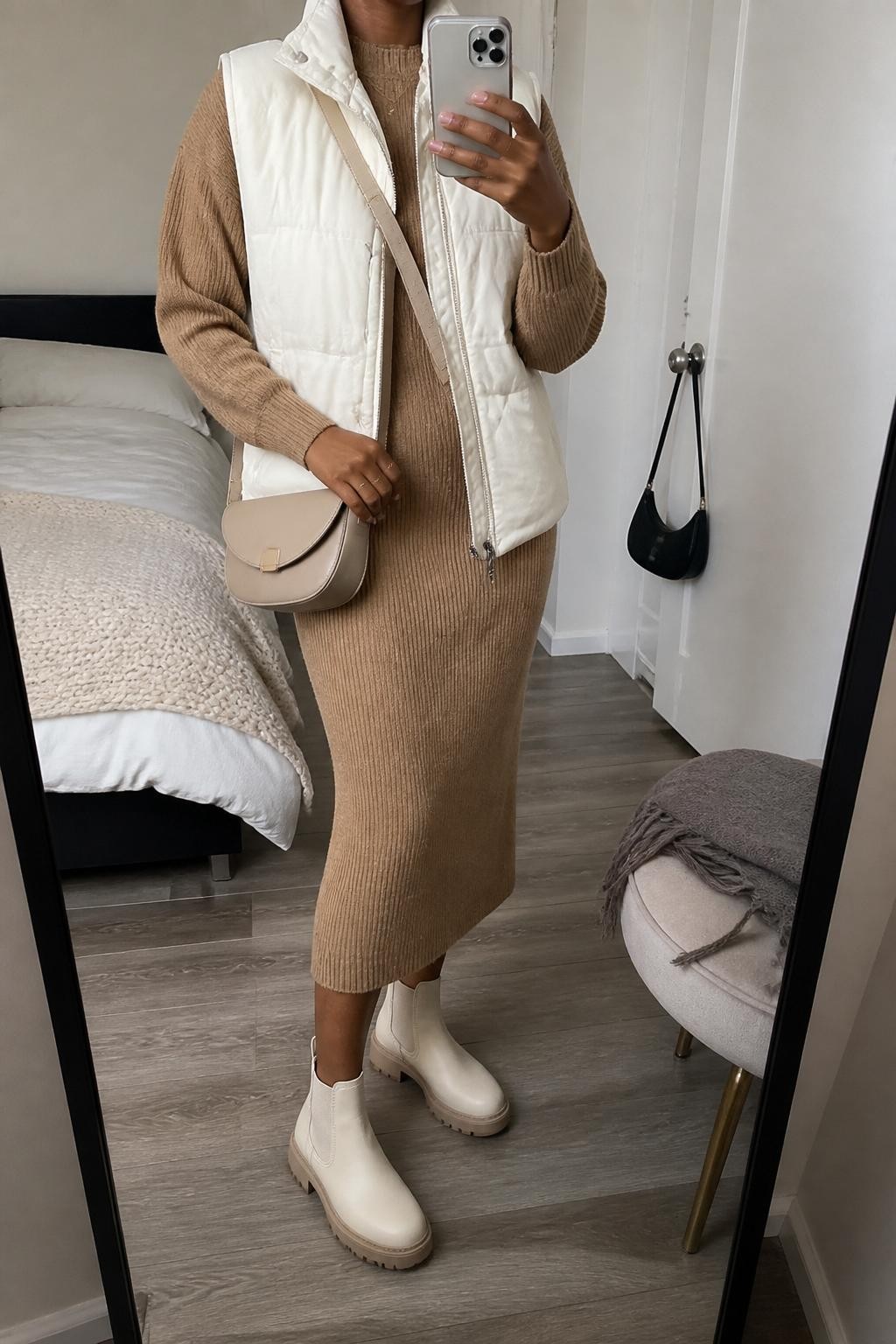 mirror selfie of a Black woman wearing the exact outfit described above, clearly showing cream Chelsea boots with slim lug soles, a camel knit sweater, matching camel knit midi skirt, an ivory quilted vest, a beige crossbody bag, and small gold studs, holding a phone in front of a mirror, cropped at NECK so the face does not show, with a relaxed casual selfie pose, in a cozy bedroom mirror setup, soft indoor lighting, with a folded blanket on a chair and a handbag hanging on a door handle, latest iPhone photo quality.