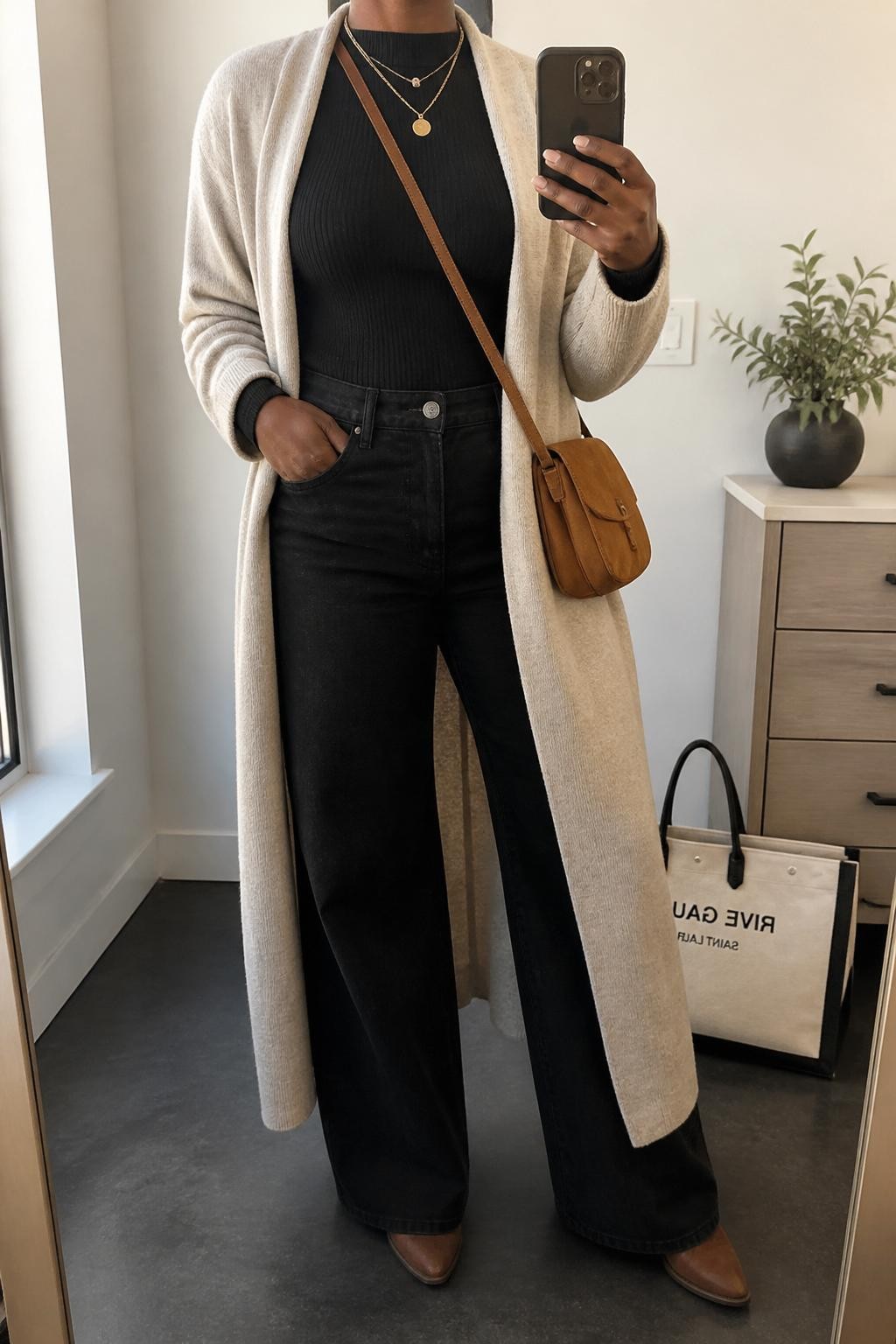 mirror selfie of a Black woman wearing the exact outfit described above, clearly showing an oatmeal long coatigan layered over a fitted black ribbed top, black wide-leg jeans, brown pointed ankle boots, a caramel suede crossbody bag, and gold layered necklaces, holding a phone in front of a mirror, cropped at NECK so the face does not show, with a relaxed casual selfie pose, in a minimal dressing room, natural window light, with a small plant on a dresser and a tote bag resting on the floor, latest iPhone photo quality.