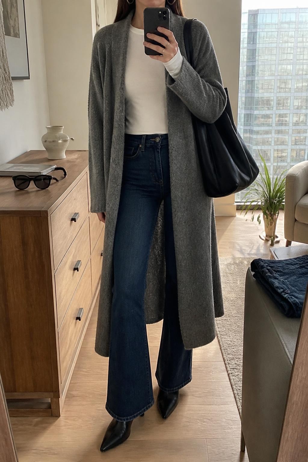 Mirror selfie of a woman wearing a long gray sweater coat, white fitted long-sleeve tee, dark wash flare jeans, black heeled ankle boots, silver hoop earrings, and a black leather tote, holding a phone in front of a mirror, cropped at NECK so the face does not show, relaxed casual selfie pose, in a stylish city apartment, natural window light, with sunglasses placed on a dresser and folded denim jeans on a bench, latest iPhone photo quality.
