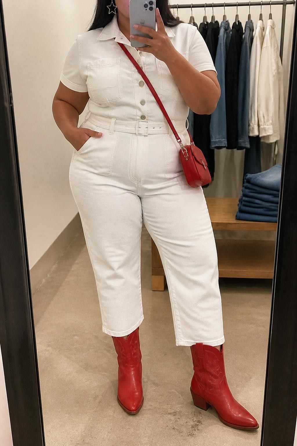 Mirror selfie of a plus size woman wearing a white denim straight-leg jumpsuit with short sleeves, silver front buttons, a fitted waist, and a matching white denim belt, bright red mid-calf cowboy boots, a small red leather crossbody bag, and silver star-shaped earrings, clearly showing the exact jumpsuit, white denim, red footwear, matching bag, buttons, and accessories listed, holding a phone in front of a mirror, cropped at the neck so the face is hidden, relaxed bold selfie pose, in a minimal dressing room with soft indoor lighting, a clothing rack with garments behind her and folded denim jeans on a bench, realistic modern vaquera styling, latest iPhone photo quality.