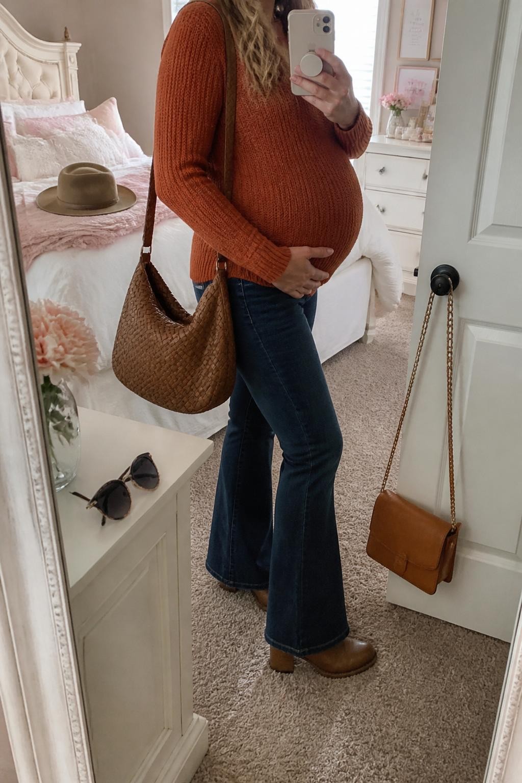 mirror selfie of a pregnant woman wearing a rust orange knit sweater, dark blue maternity flare jeans, brown heeled ankle boots, a tan wide-brim hat, and a woven brown shoulder bag, clearly showing the same clothing items, colors, fabrics, and accessories listed in the Outfit Pieces section, holding a phone in front of a mirror, cropped at NECK so the face does not show, with a relaxed casual selfie pose, in a feminine-themed bedroom, natural window light, including a handbag hanging on a door handle and sunglasses placed on a dresser, latest iPhone photo quality.