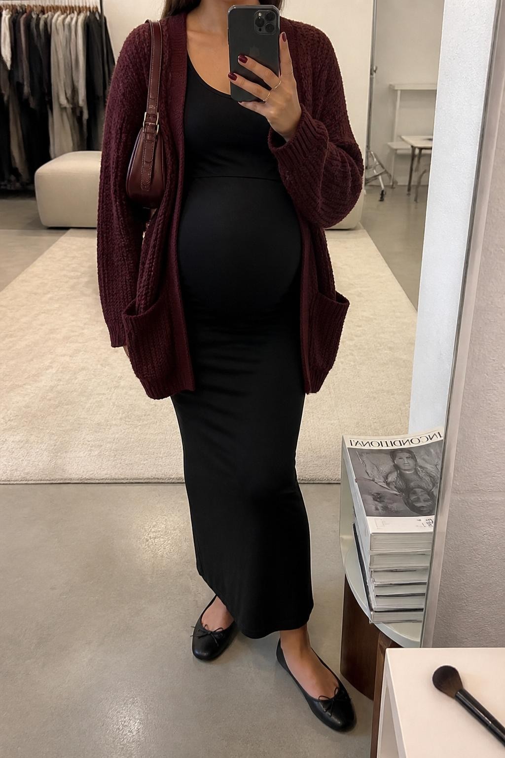 mirror selfie of a pregnant woman wearing a burgundy oversized knit cardigan, black stretchy maternity slip skirt, fitted black tank top, black ballet flats, and a burgundy shoulder bag, clearly showing the same clothing items, colors, fabrics, and accessories listed in the Outfit Pieces section, holding a phone in front of a mirror, cropped at NECK so the face does not show, with a relaxed casual selfie pose, in a fashion studio mirror corner, soft indoor lighting, including a stack of fashion magazines on a side table and a makeup brush on the counter, latest iPhone photo quality.