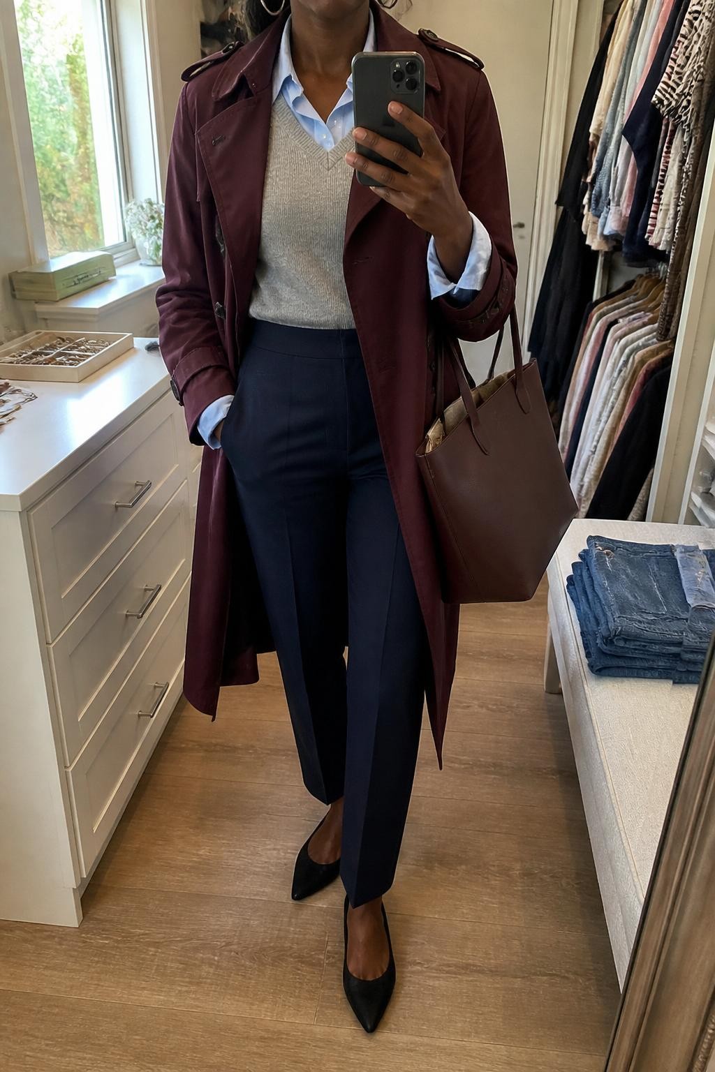 Mirror selfie of a Black woman wearing a burgundy trench coat left open over a heather gray sweater vest and a pale blue button-down shirt, clearly showing dark navy tailored trousers, black pointed-toe flats, a structured burgundy tote, and small silver hoop earrings, holding an iPhone in front of a walk-in closet mirror, cropped at the neck so the face does not show, relaxed casual selfie pose, natural window light, folded denim jeans on a bench and a jewelry tray on the vanity, natural unstaged setting, latest iPhone photo quality.