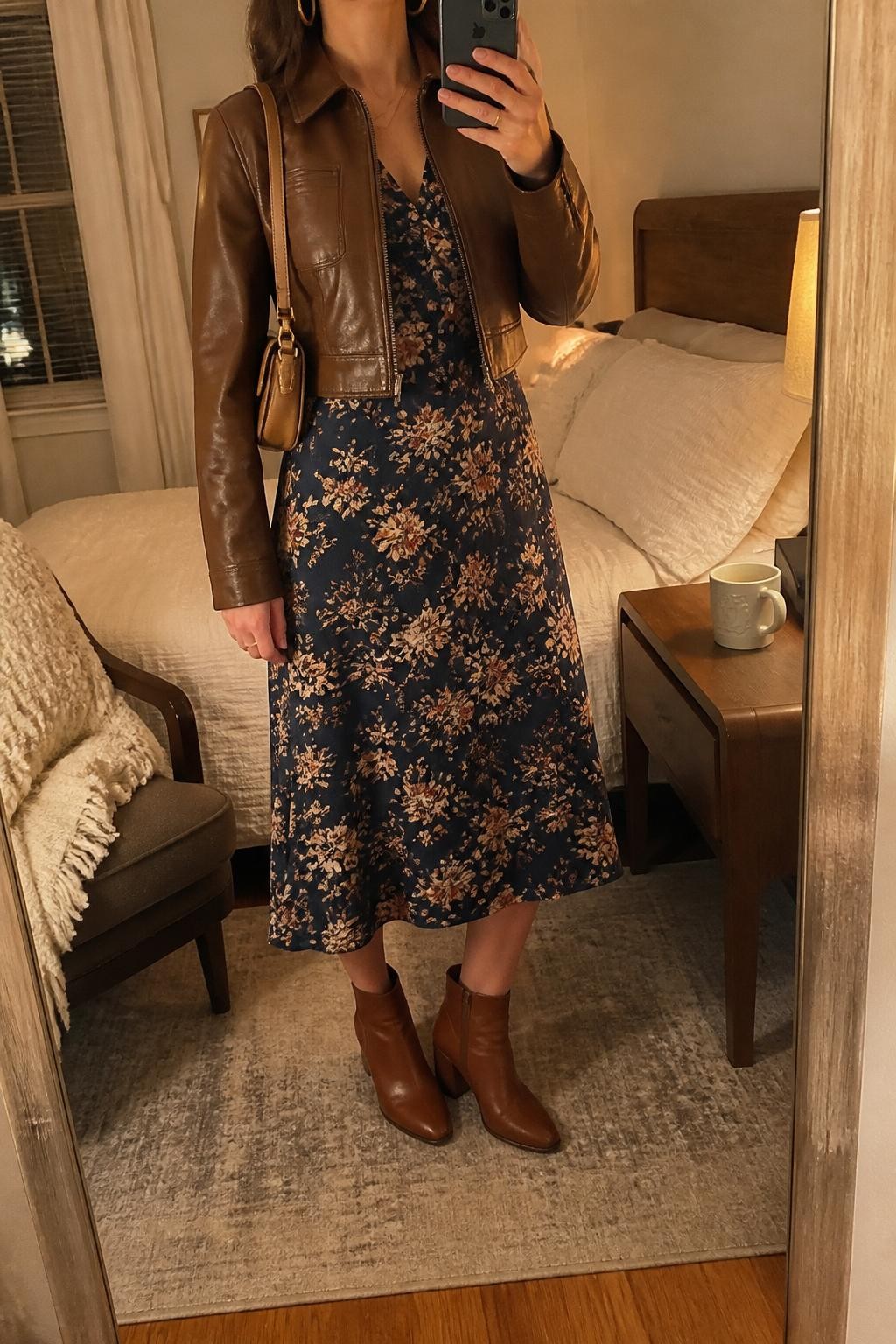 mirror selfie of a woman wearing the exact outfit described above, clearly showing a navy floral midi dress with cream and rust flowers, a cropped brown leather jacket, brown block-heel ankle boots, a tan mini shoulder bag, and gold hoop earrings, holding a phone in front of a mirror, cropped at NECK so the face does not show, with a relaxed casual selfie pose, in a cozy bedroom mirror setup, evening golden hour, with a folded blanket on a chair and a coffee mug on a bedside table, latest iPhone photo quality.