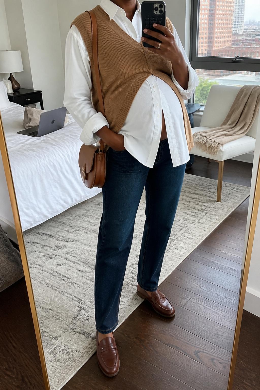 mirror selfie of a pregnant Black woman wearing a white maternity button-down shirt, camel sweater vest, dark wash maternity straight-leg jeans, brown loafers, and a tan saddle bag, clearly showing the same clothing items, colors, fabrics, and accessories listed in the Outfit Pieces section, holding a phone in front of a mirror, cropped at NECK so the face does not show, with a relaxed casual selfie pose, in a stylish city apartment, natural window light, including a laptop on the bed and a folded blanket on a chair, latest iPhone photo quality.