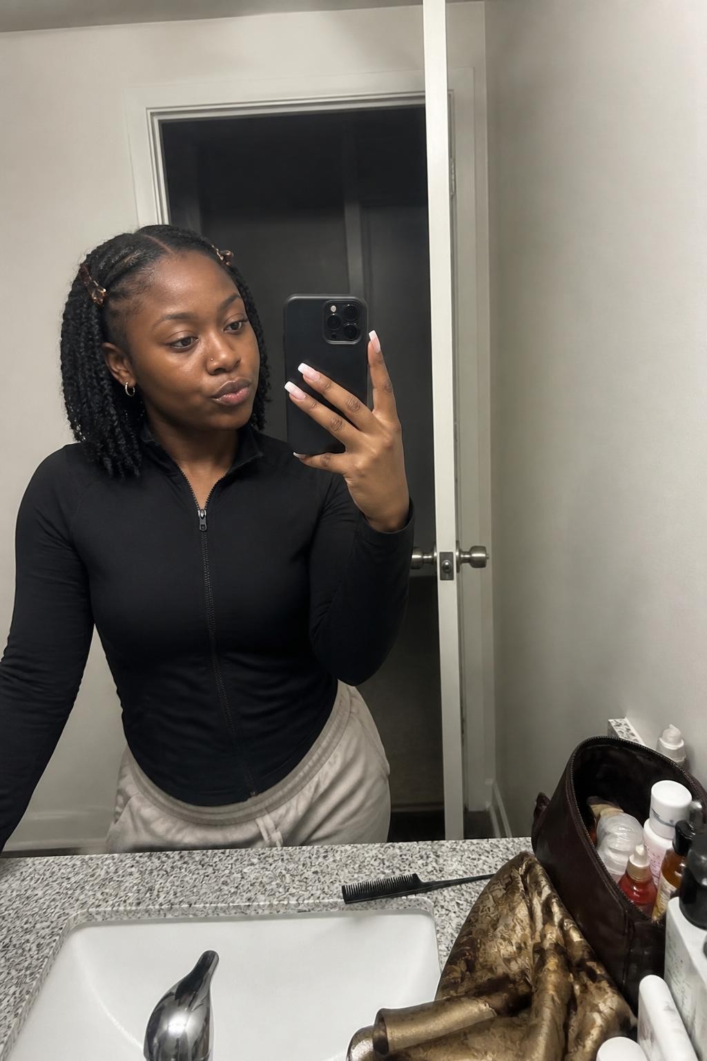 Image prompt: Photo of a Black woman wearing shoulder-length mini twists with a deep side part and small tortoiseshell clips near the front, candid mirror selfie, bathroom vanity mirror with skincare products and a makeup bag nearby, soft indoor lighting, containing a parting comb on the counter and a silk scarf draped beside the sink, realistic everyday styling moment, latest iPhone photo quality.