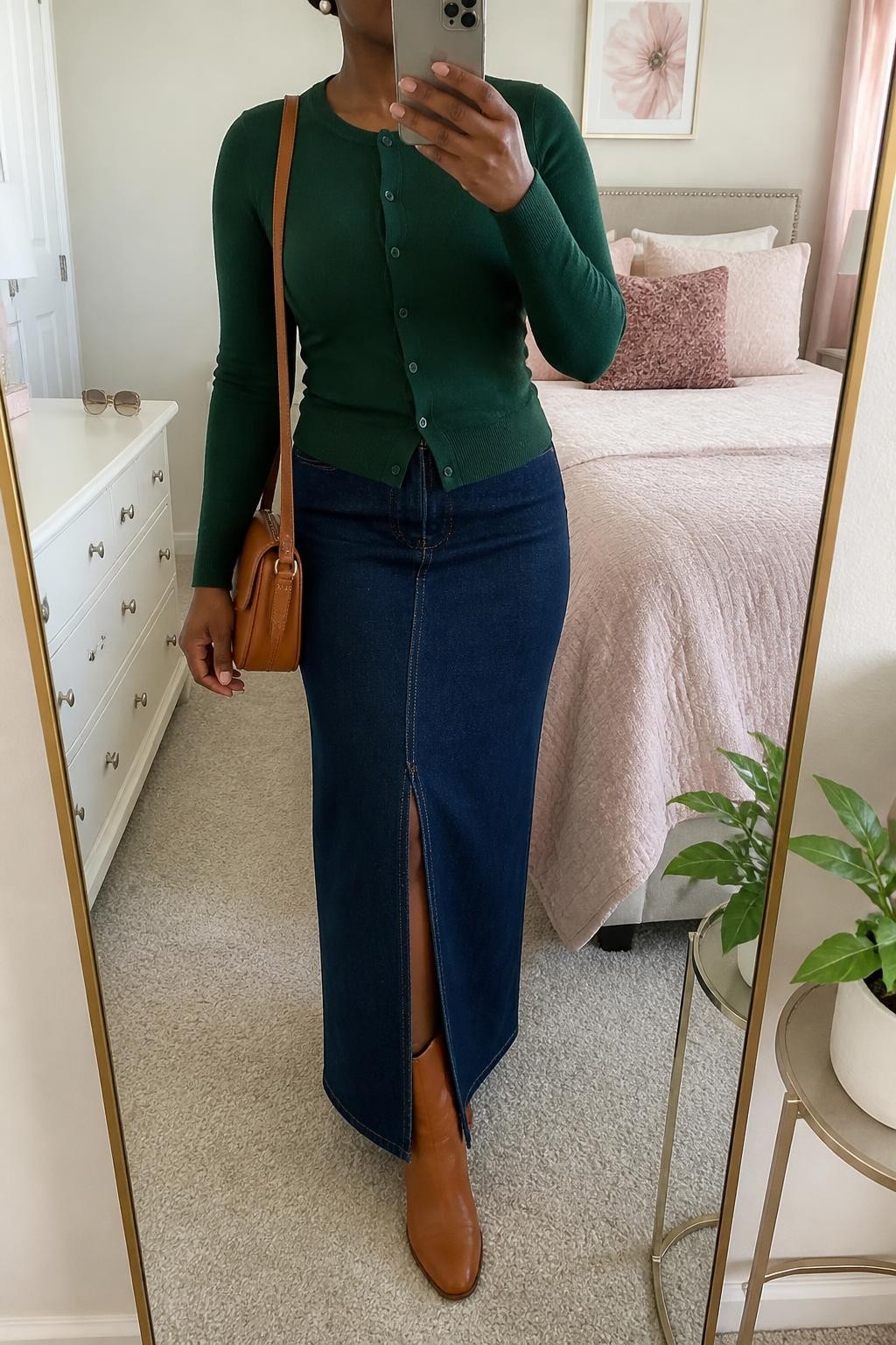 Mirror selfie of a Black woman wearing a dark indigo denim maxi skirt with a front slit, fitted forest-green button-front cardigan, tan leather ankle boots, a cognac crossbody bag, and small pearl stud earrings, clearly showing the exact clothing items, colors, fabrics, and accessories listed, holding a phone in front of a feminine-themed bedroom mirror, cropped at the neck so the face does not show, relaxed casual selfie pose, soft indoor lighting, sunglasses placed on a dresser and a small plant on a side table, realistic and uncluttered, latest iPhone photo quality.