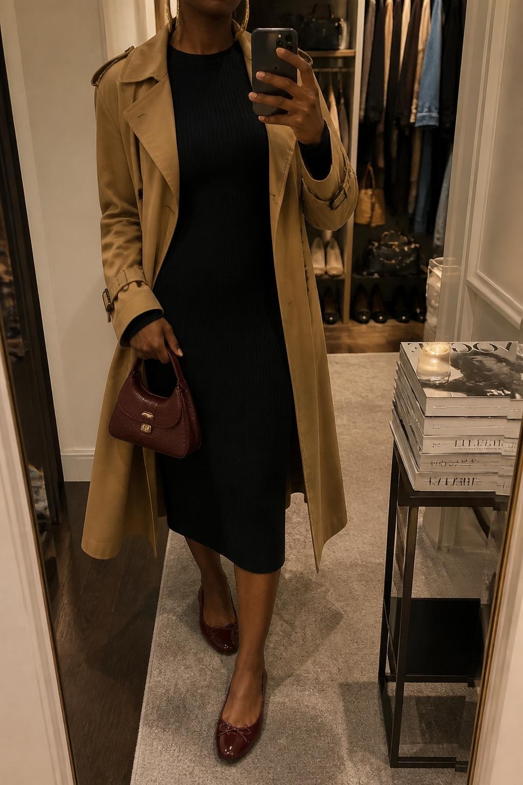 mirror selfie of a Black woman wearing the exact outfit described above, clearly showing a black ribbed long-sleeve midi dress, burgundy ballet flats, a camel trench coat worn open, a burgundy mini handbag, and gold hoop earrings, holding a phone in front of a mirror, cropped at NECK so the face does not show, with a relaxed casual selfie pose, in a walk-in closet mirror, warm moody lighting, with an open closet with clothes visible and a stack of fashion magazines on a side table, latest iPhone photo quality.