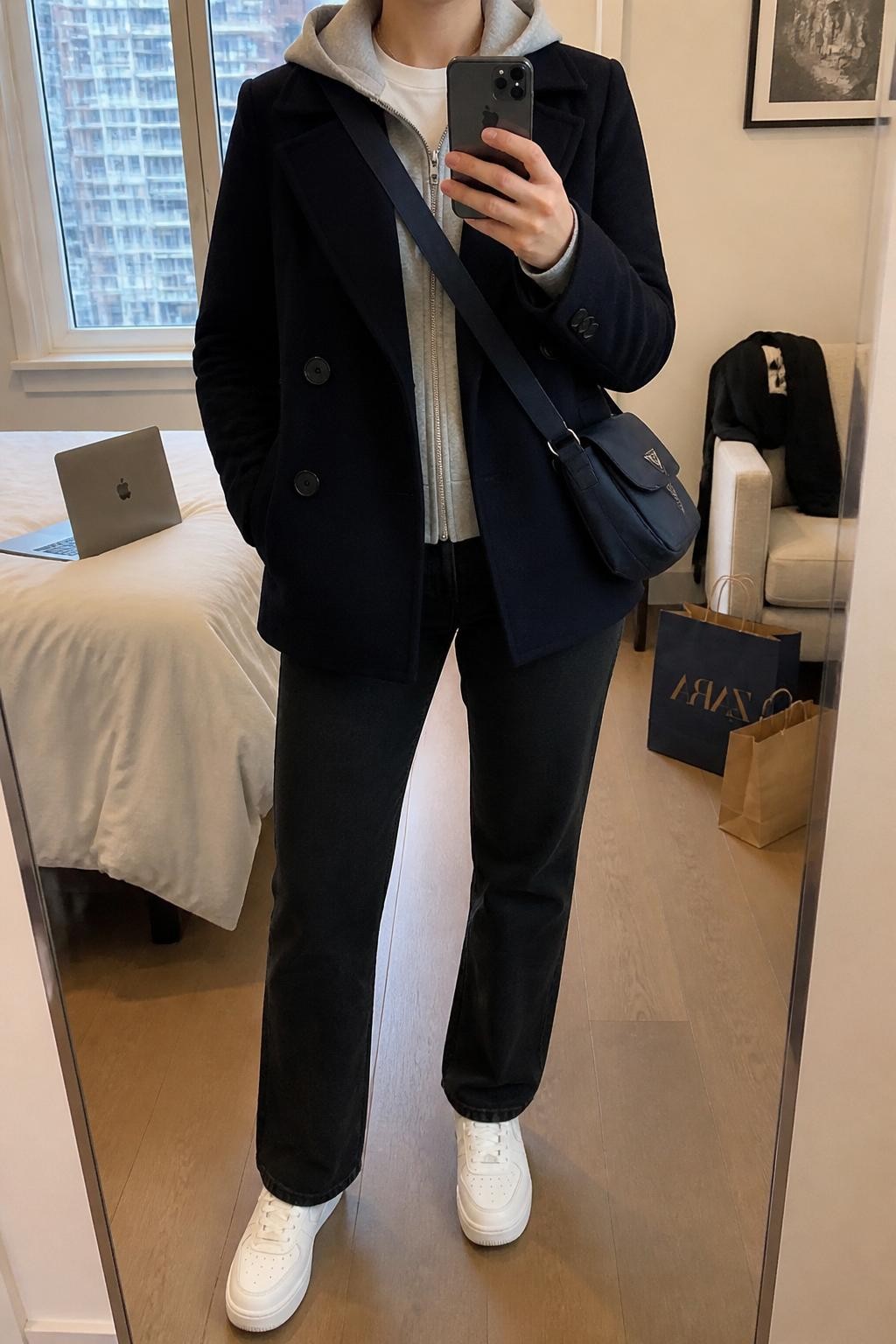 Mirror selfie of a woman wearing a structured navy peacoat over a slim heather gray hooded sweatshirt and a white crew-neck T-shirt, clearly showing black straight-leg jeans, white leather sneakers, and a navy nylon crossbody bag, holding an iPhone in front of a stylish city apartment mirror, cropped at the neck so the face does not show, relaxed casual selfie pose, soft indoor lighting, a laptop on the bed and shopping bags beside a chair, natural unstaged setting, latest iPhone photo quality.
