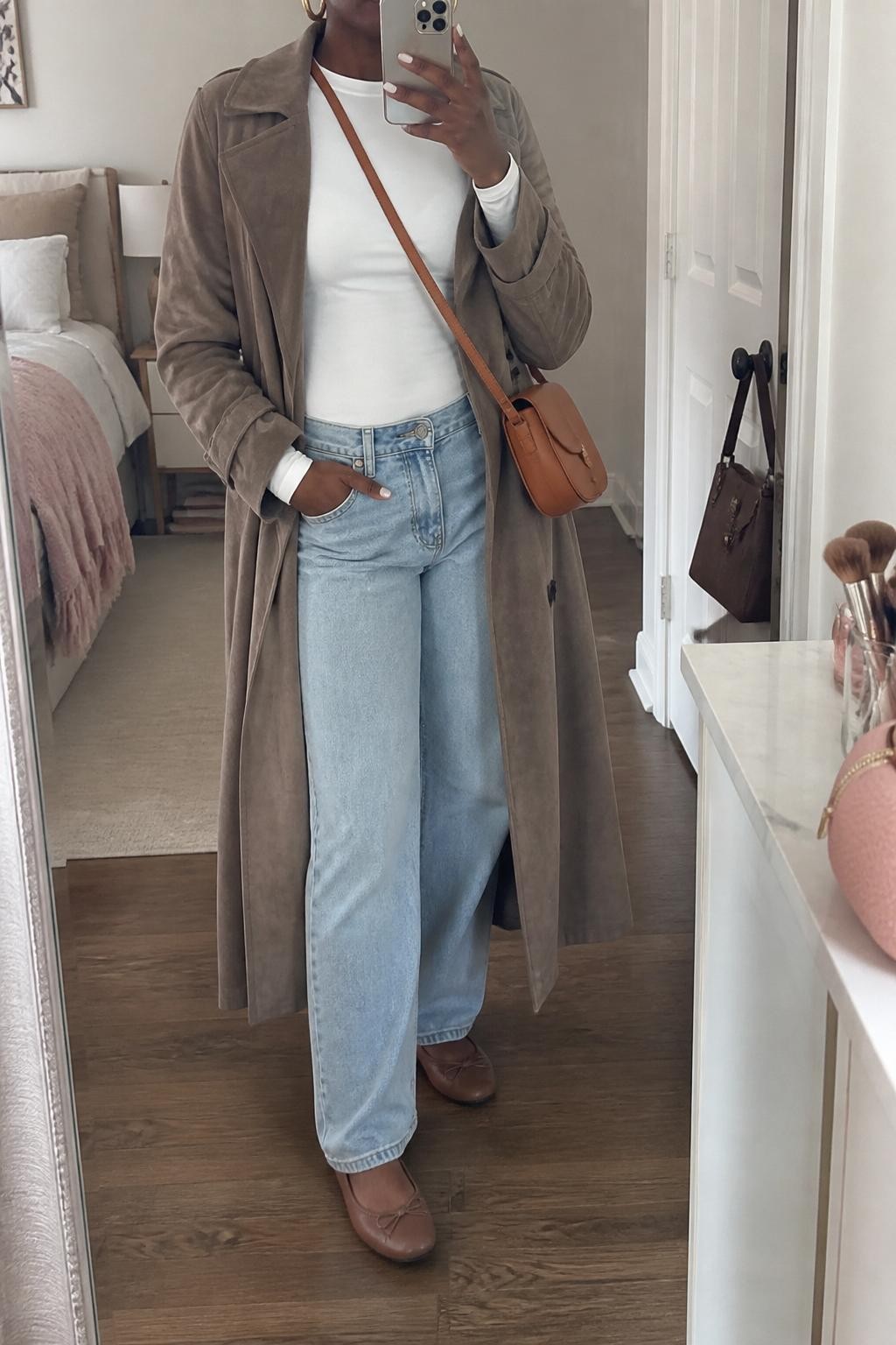 mirror selfie of a Black woman wearing the exact outfit described above, clearly showing light-wash relaxed jeans, a white fitted long-sleeve tee, a taupe suede trench coat, brown ballet flats, a tan leather crossbody bag, and gold hoop earrings, holding a phone in front of a mirror, cropped at NECK so the face does not show, with a relaxed casual selfie pose, in a feminine-themed bedroom, natural window light, with a makeup brush or cosmetic bag on the counter and a handbag hanging on a door handle, latest iPhone photo quality.
