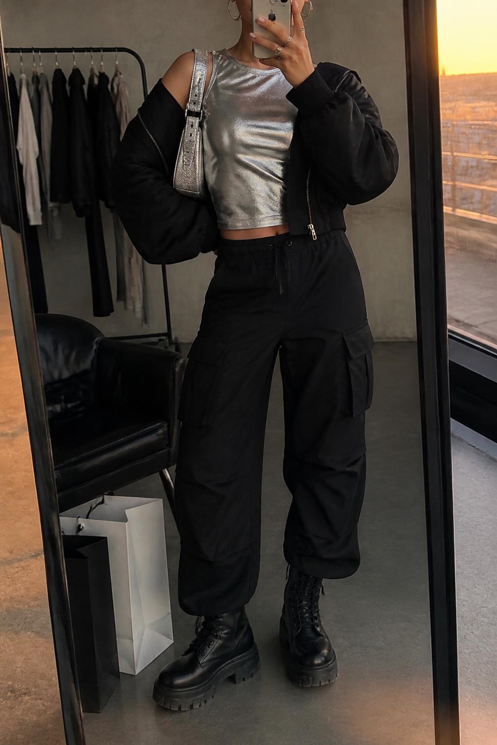 mirror selfie of a woman wearing the exact outfit described above, clearly showing a silver metallic sleeveless top, black relaxed cargo pants, black platform combat boots, a black cropped bomber jacket, a silver mini shoulder bag, and small silver hoop earrings, holding a phone in front of a mirror, cropped at NECK so the face does not show, with a relaxed casual selfie pose, in a fashion studio mirror corner, evening golden hour, with a clothing rack with garments behind and shopping bags beside a chair, latest iPhone photo quality.