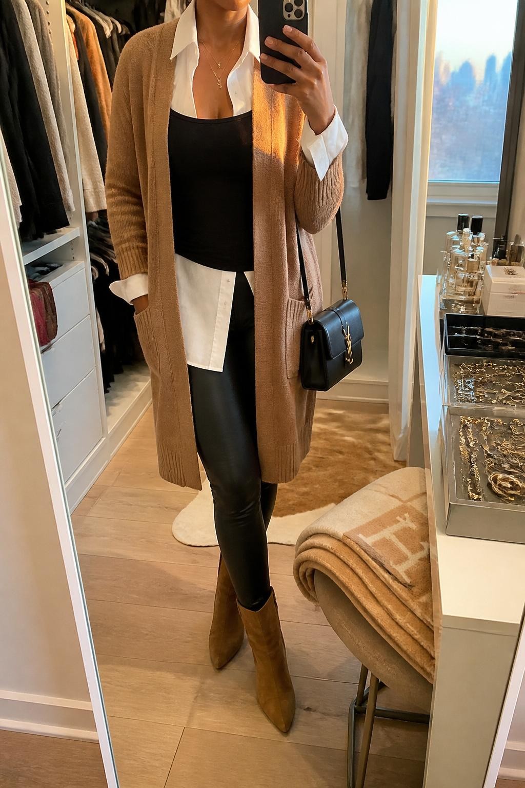 Mirror selfie of a Black woman wearing a camel longline cardigan over a crisp white poplin shirt and a fitted black camisole, clearly showing black leather leggings, camel suede ankle boots, and a structured black shoulder bag, holding an iPhone in front of a walk-in closet mirror, cropped at the neck so the face does not show, relaxed casual selfie pose, evening golden hour light, a jewelry tray on the vanity and a folded blanket on a chair, natural unstaged setting, latest iPhone photo quality.