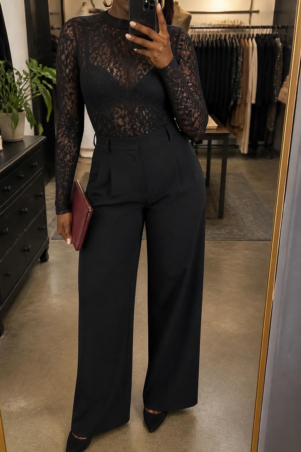mirror selfie of a Black woman wearing the exact outfit described above, clearly showing a black lined lace top with long sleeves, black high-waisted wide-leg trousers, black suede pumps, a burgundy clutch, and gold hoop earrings, holding a phone in front of a mirror, cropped at NECK so the face does not show, with a relaxed casual selfie pose, in a fashion studio mirror corner, warm moody lighting, with a clothing rack with garments behind and a small plant on a dresser, latest iPhone photo quality.