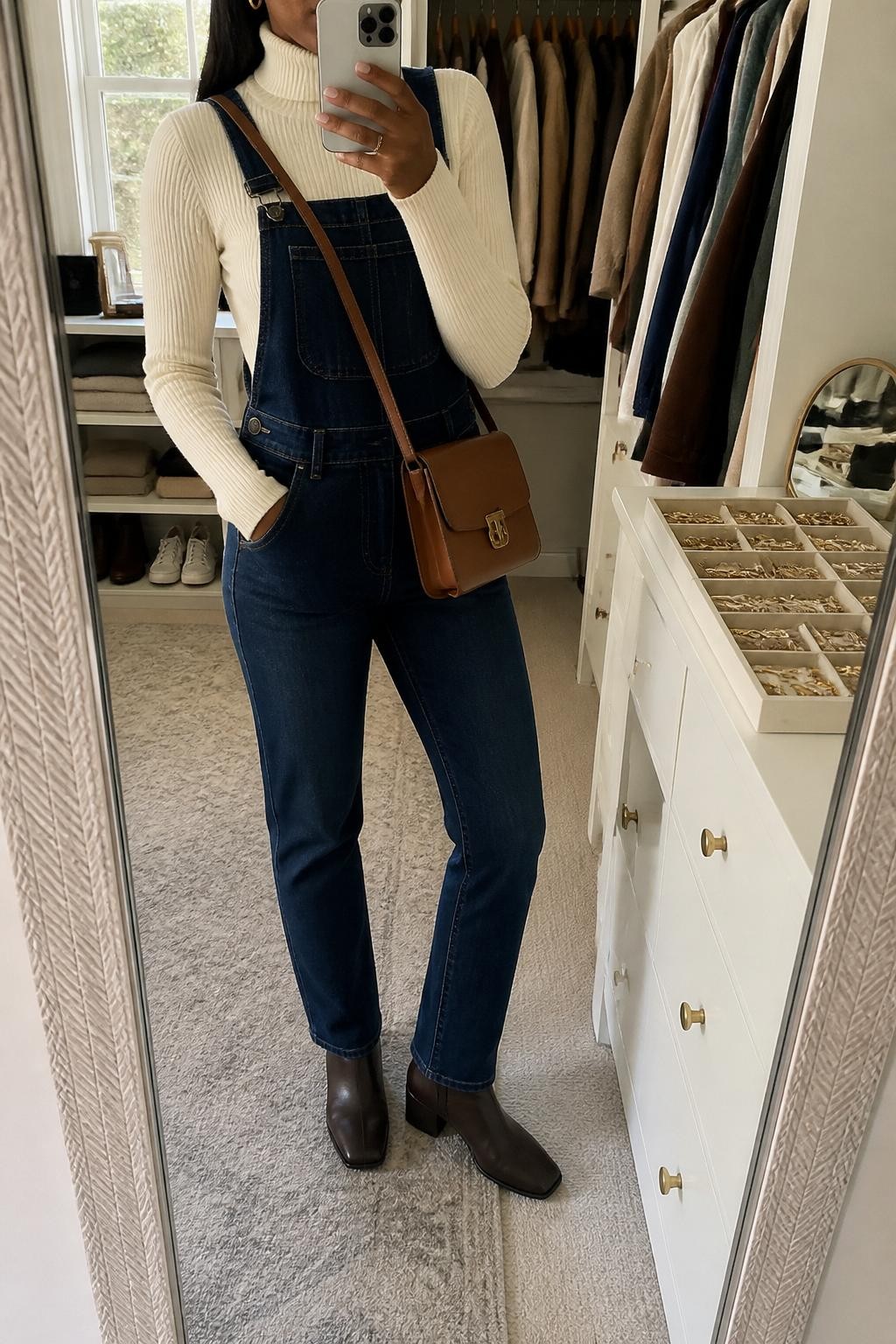 Mirror selfie of a Black woman wearing fitted dark indigo straight-leg denim overalls over a cream ribbed turtleneck, clearly showing dark brown leather ankle boots, a structured cognac crossbody bag, and small gold hoop earrings, holding an iPhone in front of a walk-in closet mirror, cropped at the neck so the face does not show, relaxed casual selfie pose, natural window light, open closet with clothes visible and a jewelry tray on the vanity, natural unstaged setting, latest iPhone photo quality.
