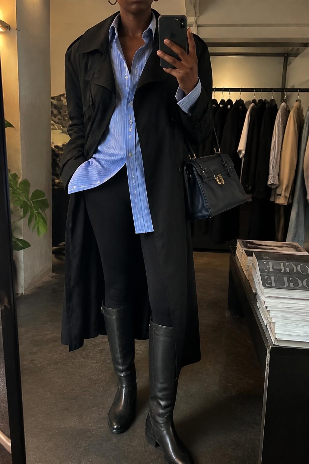 Mirror selfie of a Black woman wearing a long black trench coat over an oversized blue-and-white striped button-down shirt and fitted black leggings, clearly showing tall black riding boots, a structured dark navy handbag, and small silver hoop earrings, holding an iPhone in front of a fashion studio mirror corner, cropped at the neck so the face does not show, relaxed casual selfie pose, warm moody lighting, a clothing rack with garments behind and a stack of fashion magazines on a side table, natural unstaged setting, latest iPhone photo quality.