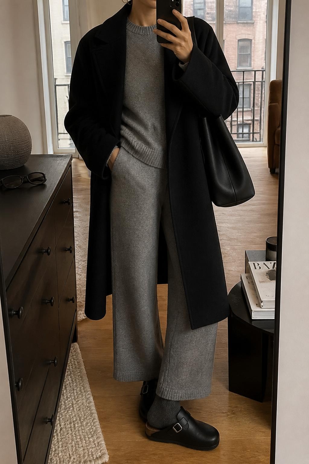 mirror selfie of a woman wearing the exact outfit described above, clearly showing black leather Birkenstock-style clogs with charcoal socks, a soft gray knit sweater, matching gray knit wide-leg pants, a black wool coat worn open, a black leather tote bag, and silver hoop earrings, holding a phone in front of a mirror, cropped at NECK so the face does not show, with a relaxed casual selfie pose, in a stylish city apartment, warm moody lighting, with sunglasses placed on a dresser and a stack of fashion magazines on a side table, latest iPhone photo quality.