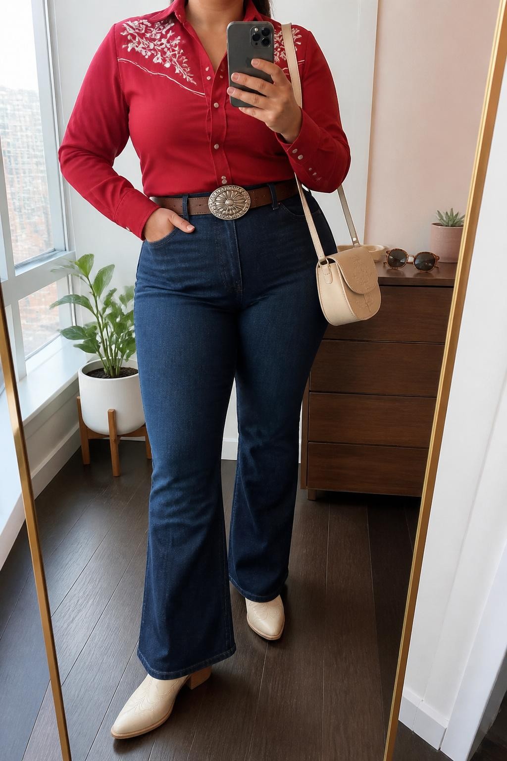 Mirror selfie of a plus size woman wearing dark indigo high-waisted flared jeans, a fitted red Western shirt with cream floral embroidery across the shoulders and silver snap buttons, pointed cream leather cowboy boots, a dark brown leather belt with a large silver oval buckle, and a small cream saddle bag, clearly showing the exact clothing items, colors, embroidery, denim wash, footwear, belt, and accessories listed, holding a phone in front of a mirror, cropped at the neck so the face does not show, relaxed confident selfie pose, in a stylish city apartment with natural window light, sunglasses placed on a dresser and a small plant nearby, natural unstaged appearance, latest iPhone photo quality.