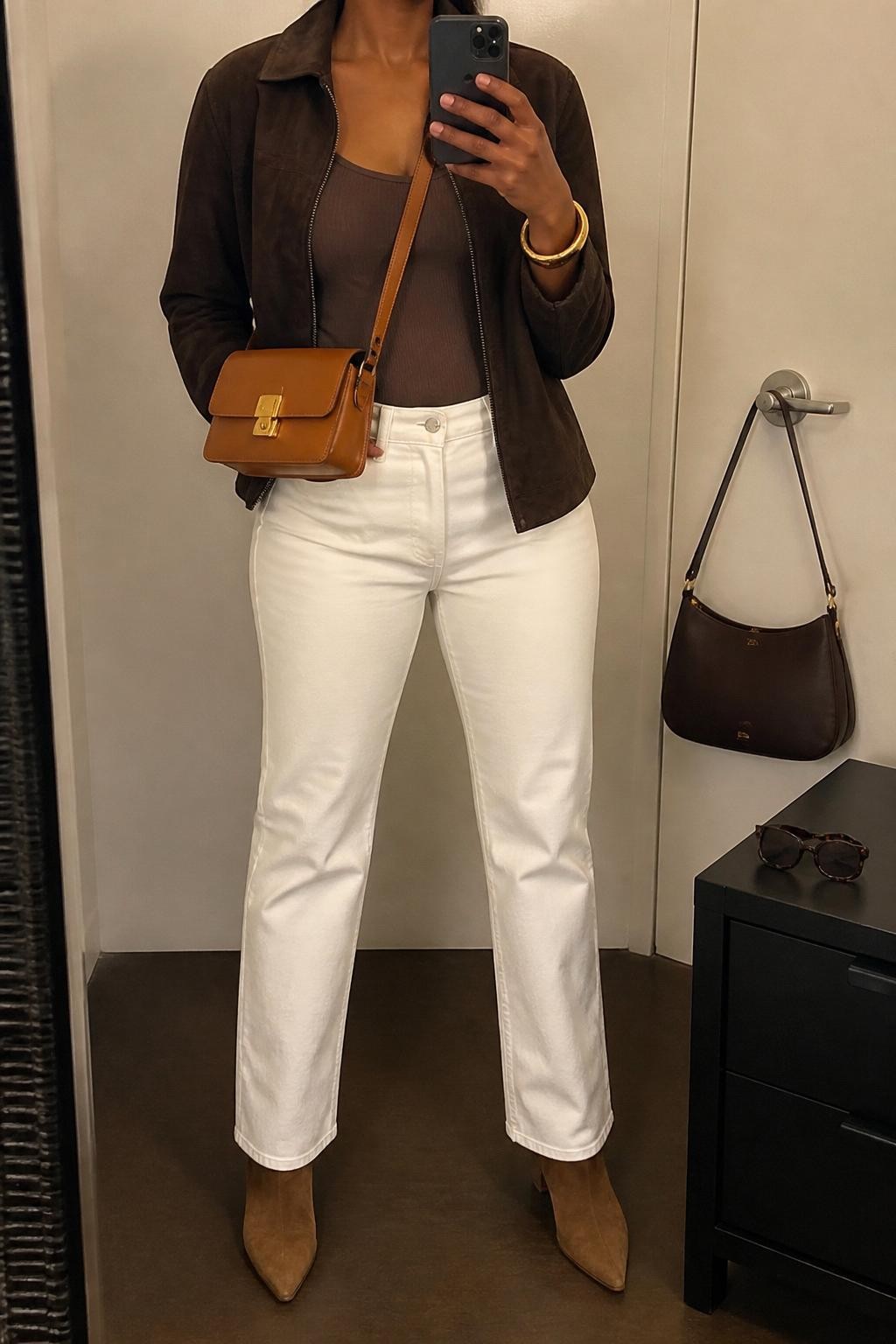 mirror selfie of a Black woman wearing the exact outfit described above, clearly showing white straight-leg jeans, a mocha fitted knit top, a chocolate brown suede jacket, tan ankle boots, a caramel leather crossbody bag, and gold bangles, holding a phone in front of a mirror, cropped at NECK so the face does not show, with a relaxed casual selfie pose, in a minimal dressing room, warm moody lighting, with a handbag hanging on a door handle and sunglasses placed on a dresser, latest iPhone photo quality.