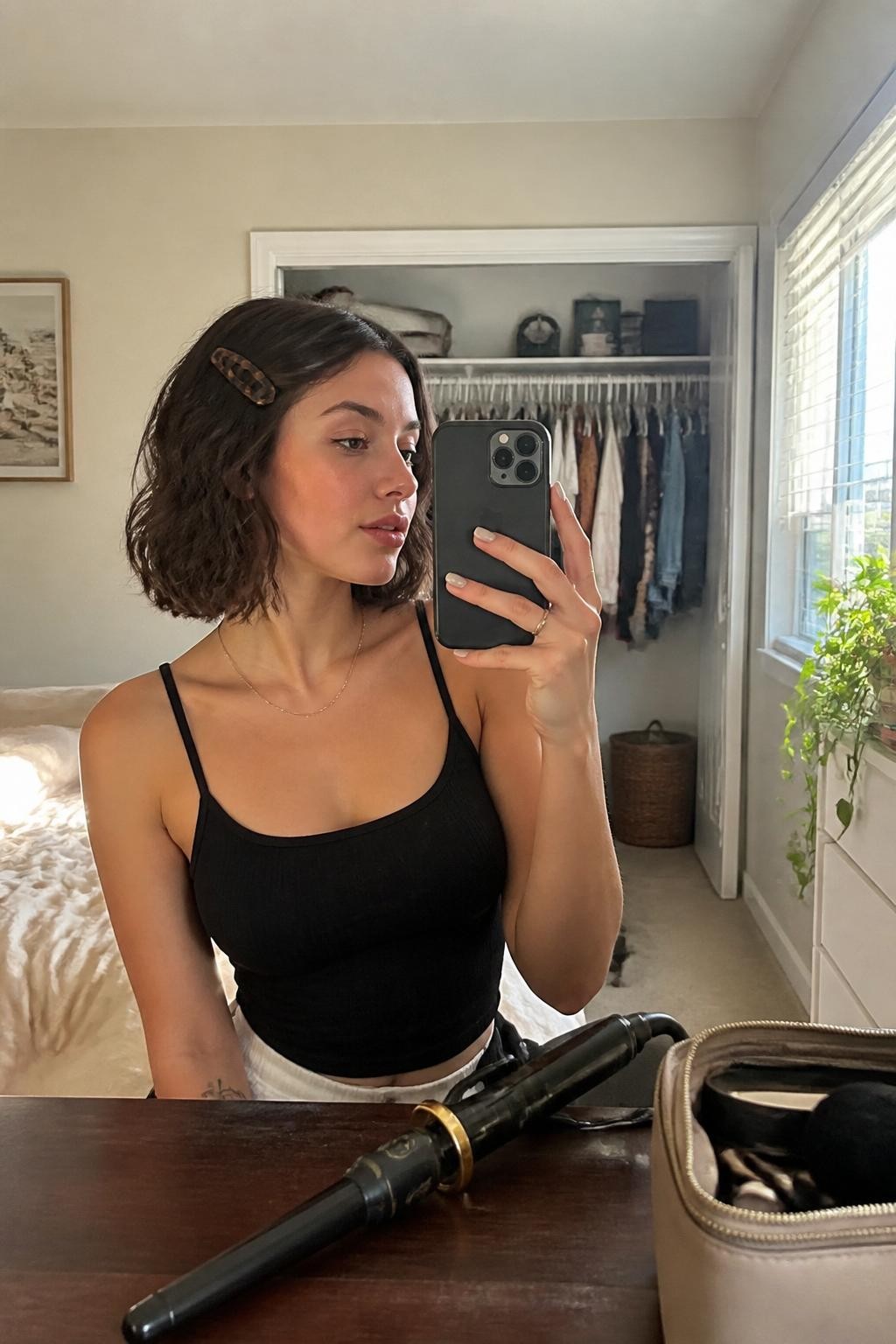 Image prompt: Photo of a woman wearing a softly waved chin-length bob with one side secured by a tortoiseshell barrette, candid mirror selfie, sun-drenched bedroom with a slightly open closet behind her, natural window light, containing a curling wand on the dresser and a makeup bag nearby, realistic morning styling moment, latest iPhone photo quality.