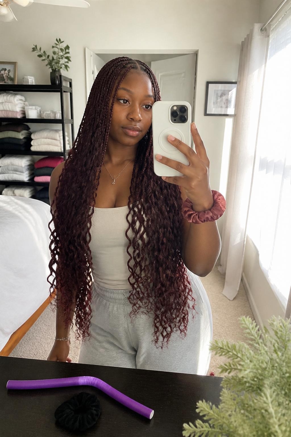 Image prompt: Photo of a Black woman wearing waist-length burgundy boho braids with loose curls woven throughout and a clean center part, candid mirror selfie, sun-drenched bedroom with folded sweaters visible behind her, natural window light, containing a flexi rod on the dresser and a silk scrunchie around her wrist, realistic home getting-ready moment, latest iPhone photo quality.