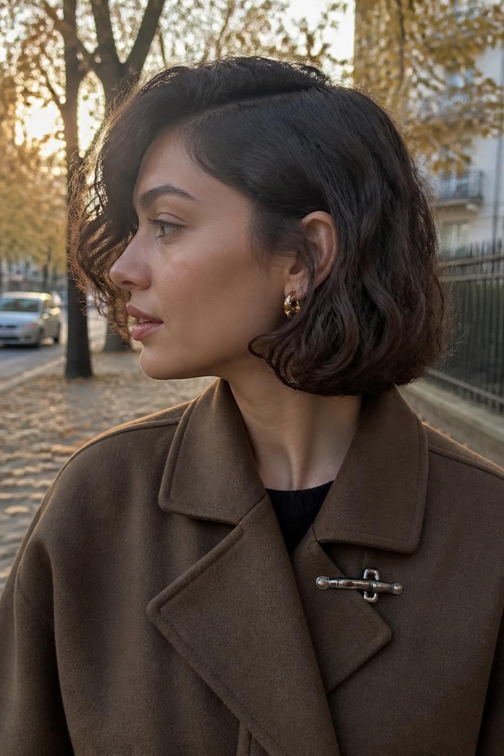 Image prompt: Photo of a woman wearing a chin-length curly bob with a deep side part, soft rounded volume, and one side tucked behind her ear, side profile view, softly blurred city street background with fallen autumn leaves, golden hour glow, containing small gold statement earrings peeking through and a hair clip attached to her coat collar, realistic casual moment, latest iPhone photo quality.