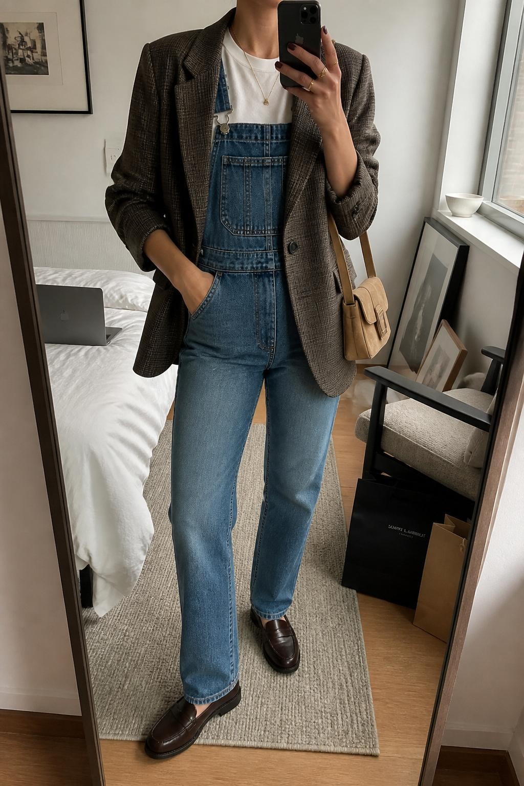 Mirror selfie of a woman wearing medium-wash straight-leg denim overalls over a fitted white crew-neck T-shirt with a tailored brown plaid blazer layered on top, clearly showing dark brown leather loafers and a tan rectangular shoulder bag, holding an iPhone in front of a stylish city apartment mirror, cropped at the neck so the face does not show, relaxed casual selfie pose, natural window light, a laptop on the bed and shopping bags beside a chair, natural unstaged setting, latest iPhone photo quality.