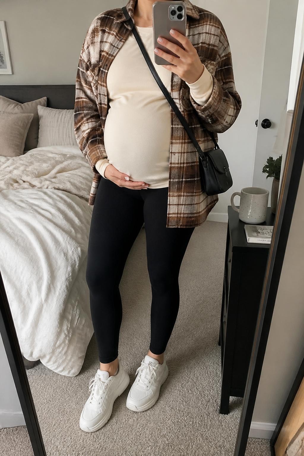 mirror selfie of a pregnant woman wearing black maternity leggings, an ivory long sleeve maternity tee, an oversized brown and cream plaid shacket, white chunky sneakers, and a black crossbody bag, clearly showing the same clothing items, colors, fabrics, and accessories listed in the Outfit Pieces section, holding a phone in front of a mirror, cropped at NECK so the face does not show, with a relaxed casual selfie pose, in a cozy bedroom mirror setup, soft indoor lighting, including soft pillows on an unmade bed and a coffee mug on a bedside table, latest iPhone photo quality.