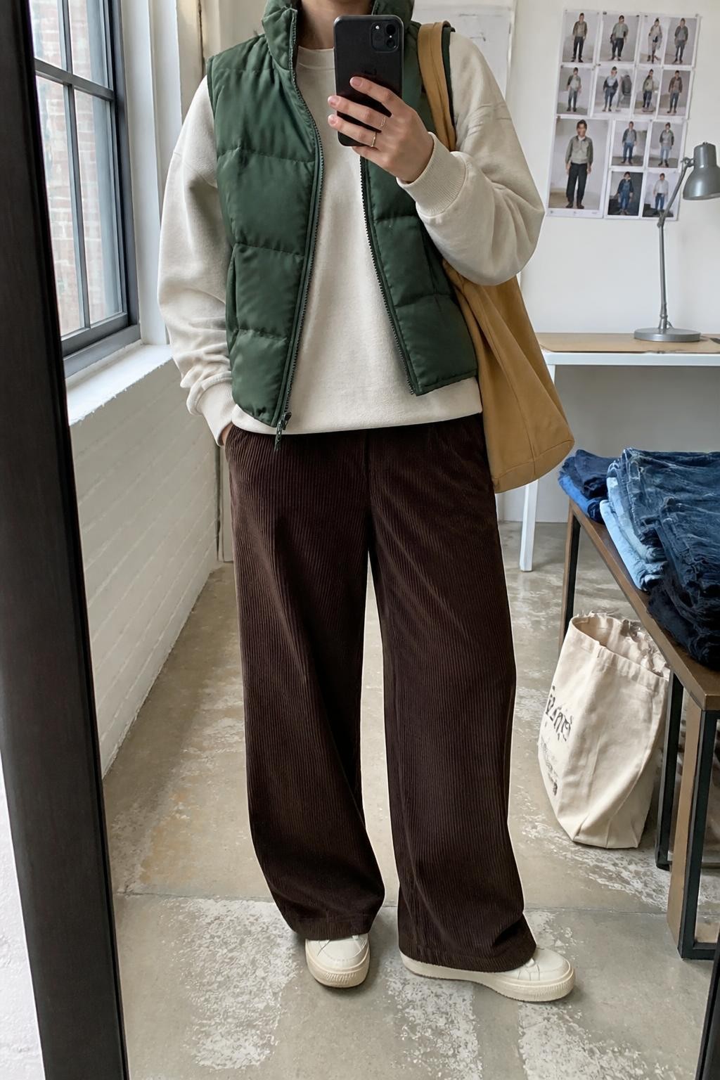 Mirror selfie of a woman wearing a forest-green quilted puffer vest over an oversized oatmeal crew-neck sweatshirt, chocolate-brown wide-leg corduroy pants, cream leather sneakers, and a tan canvas tote bag, clearly showing the exact clothing items, colors, fabrics, and accessories listed, holding a phone in front of a fashion studio mirror corner, cropped at the neck so the face does not show, relaxed casual selfie pose, natural window light, folded denim jeans on a bench and a tote bag resting on the floor, casual realistic setting, latest iPhone photo quality.