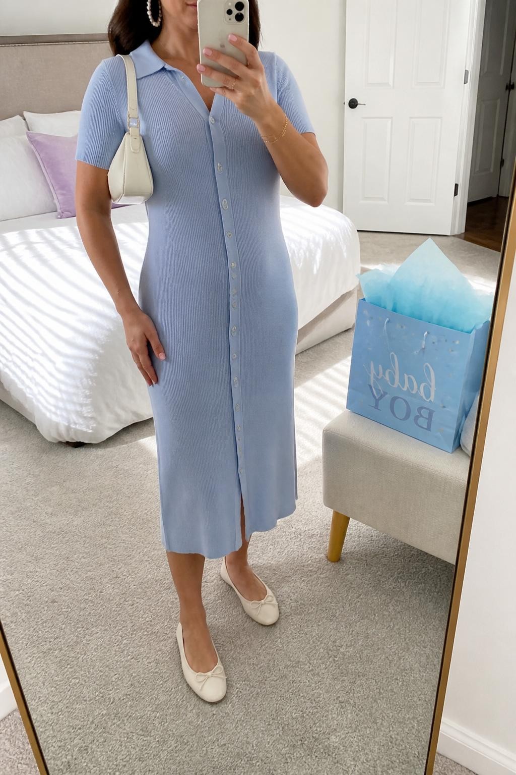 mirror selfie of a woman wearing the exact outfit described above, clearly showing a soft blue button-front cardigan midi dress, cream ballet flats, a white mini shoulder bag, pearl hoop earrings, and a delicate gold bracelet, holding a phone in front of a mirror, cropped at NECK so the face does not show, with a relaxed casual selfie pose, in a bright bedroom mirror setup, natural daylight, with a blue baby shower gift bag on a chair and tissue paper peeking out, latest iPhone photo quality.
