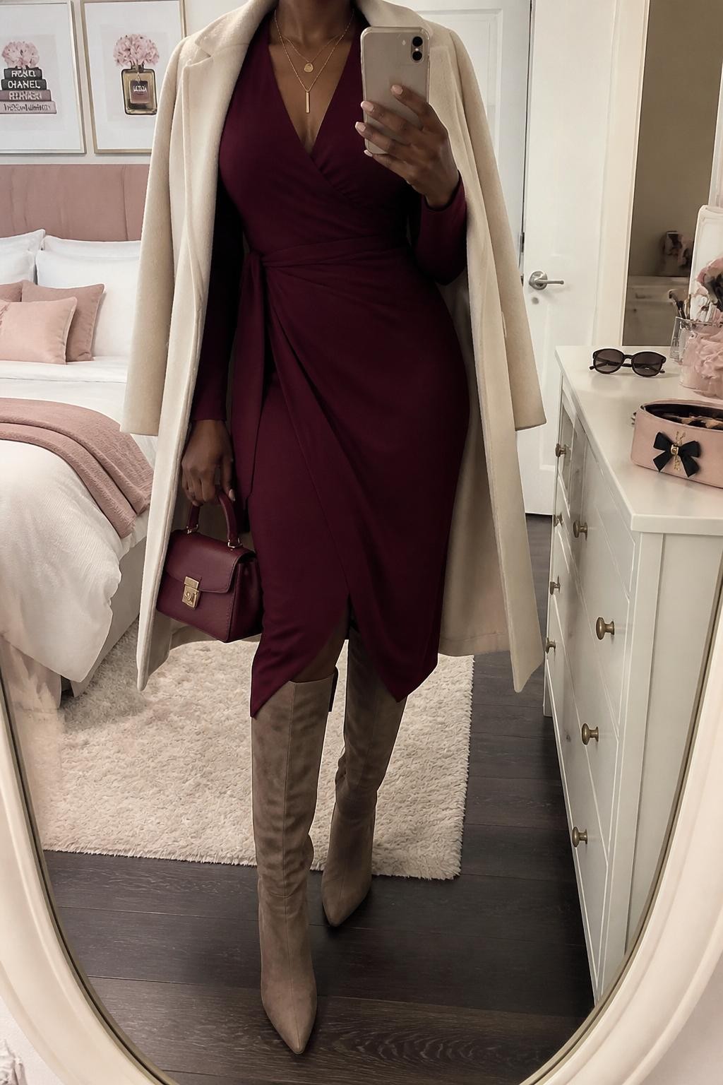 mirror selfie of a Black woman wearing the exact outfit described above, clearly showing a burgundy long-sleeve wrap midi dress, taupe suede knee-high boots, a cream wool coat draped open, a burgundy mini handbag, and delicate gold layered necklaces, holding a phone in front of a mirror, cropped at NECK so the face does not show, with a relaxed casual selfie pose, in a feminine-themed bedroom, soft indoor lighting, with sunglasses placed on a dresser and a makeup brush or cosmetic bag on the counter, latest iPhone photo quality.