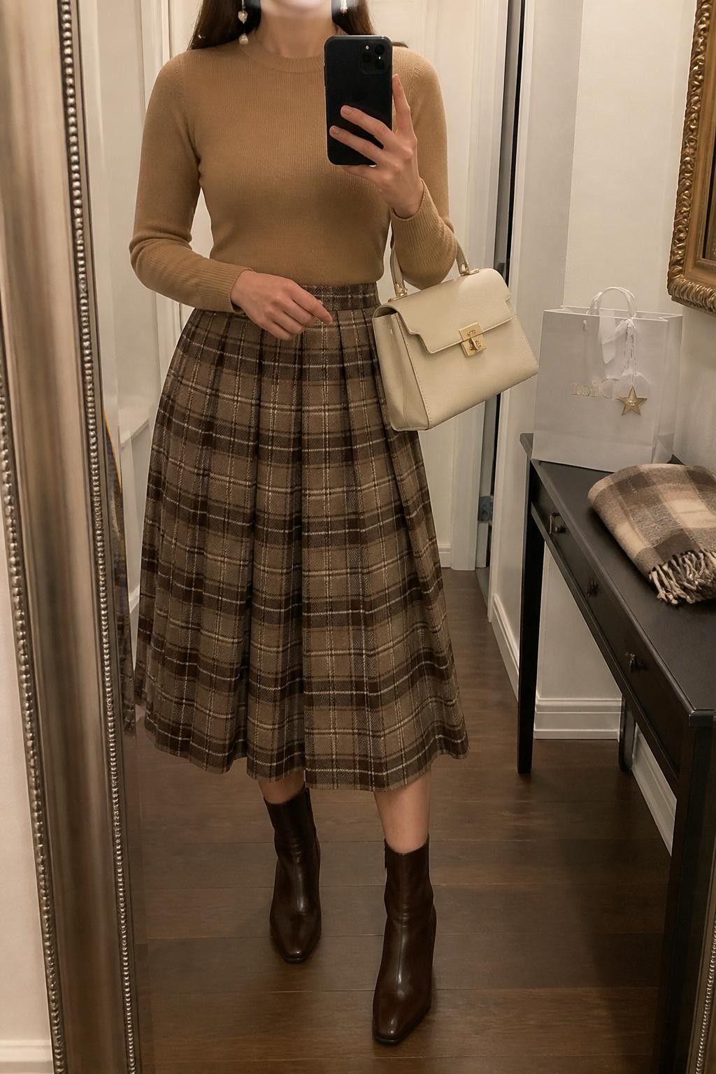 mirror selfie of a woman wearing the exact outfit described above, clearly showing a fitted camel knit top tucked into a brown and cream plaid pleated midi skirt, dark brown ankle boots, a cream top-handle bag, and pearl drop earrings, holding a phone in front of a mirror, cropped at NECK so the face does not show, with a relaxed casual selfie pose, in a feminine apartment hallway mirror, warm indoor lighting, with a gift bag on a console table and a folded scarf beside it, latest iPhone photo quality.
