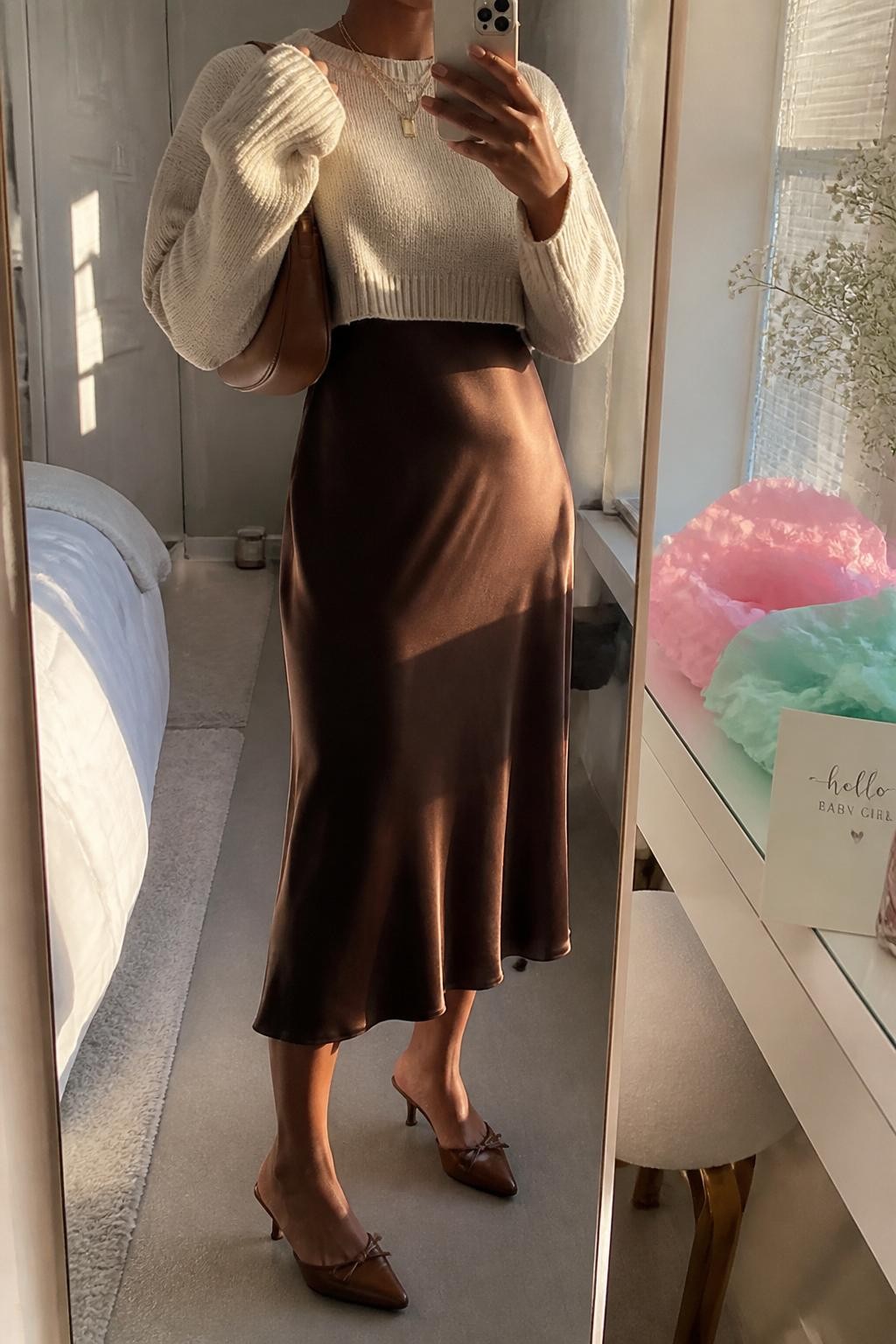 mirror selfie of a Black woman wearing the exact outfit described above, clearly showing a chocolate brown satin midi slip dress layered under an oatmeal cropped knit sweater, brown kitten-heel mules, a tan crescent shoulder bag, and gold layered necklaces, holding a phone in front of a mirror, cropped at NECK so the face does not show, with a relaxed casual selfie pose, in a modern bedroom mirror setup, soft golden hour light, with pastel tissue paper and a baby shower card on the vanity, latest iPhone photo quality.