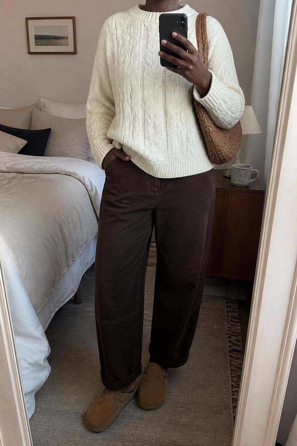 mirror selfie of a Black woman wearing the exact outfit described above, clearly showing chocolate brown barrel jeans, a cream cable knit sweater, tan suede clogs, a caramel woven shoulder bag, and small gold stud earrings, holding a phone in front of a mirror, cropped at NECK so the face does not show, with a relaxed casual selfie pose, in a cozy bedroom mirror setup, soft indoor lighting, with a coffee mug on a bedside table and soft pillows on an unmade bed, latest iPhone photo quality.