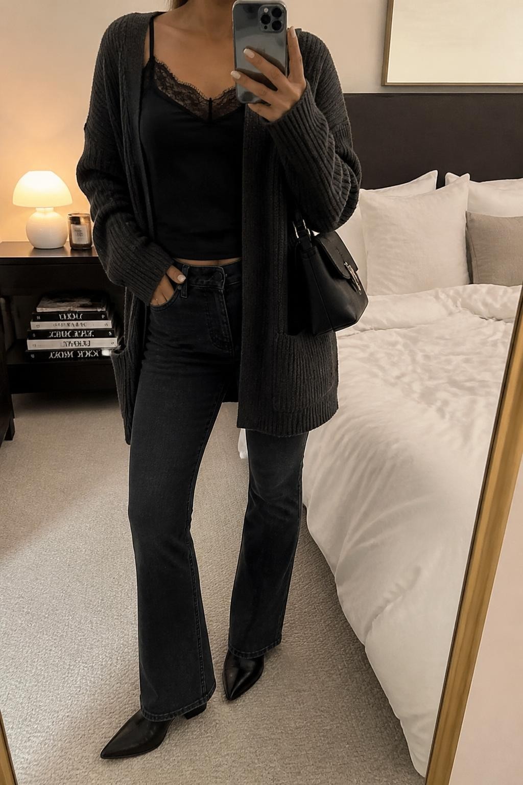 mirror selfie of a woman wearing a black lace-trim cami, charcoal longline cardigan, washed black bootcut jeans, black pointed ankle boots, and a small black satin shoulder bag, clearly showing the same clothing items, colors, fabrics, and accessories listed in the Outfit Pieces section, holding a phone in front of a mirror, cropped at NECK so the face does not show, with a relaxed casual selfie pose, in a cozy bedroom mirror setup, soft indoor lighting, including soft pillows on an unmade bed and a stack of fashion magazines on a side table, latest iPhone photo quality.