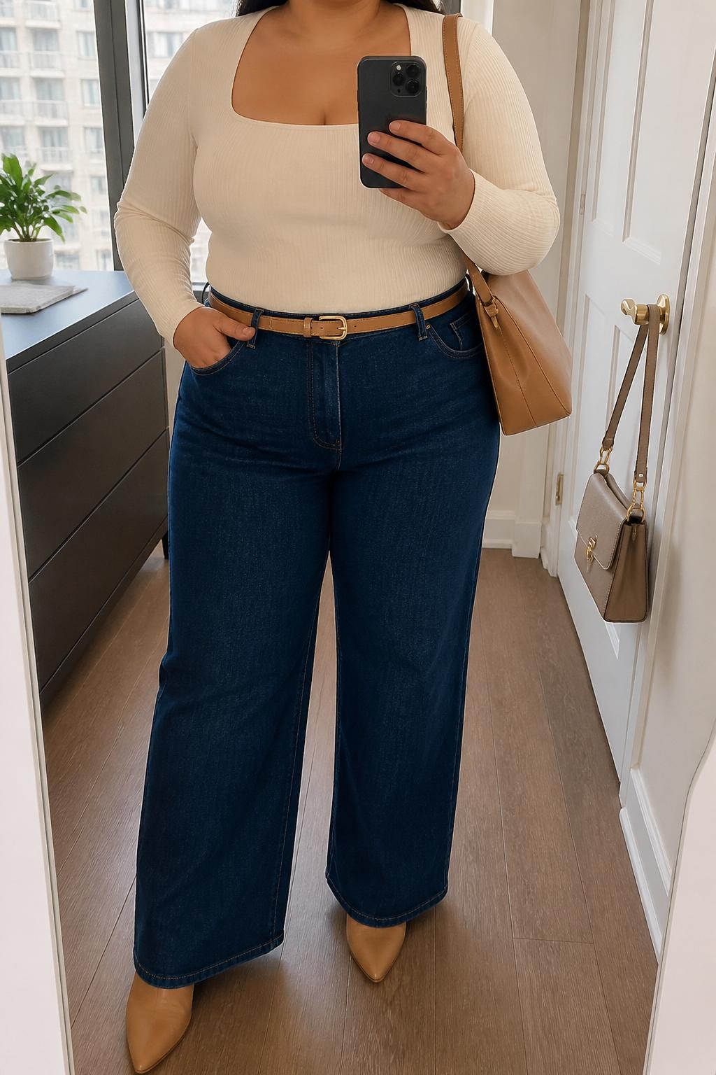 Mirror selfie of a plus size woman wearing dark indigo high-waisted wide-leg jeans, a fitted cream ribbed square-neck bodysuit, a slim camel leather belt, pointed tan ankle boots, and a structured camel shoulder bag, clearly showing the exact clothing items, colors, denim wash, ribbed texture, footwear, belt, and accessories listed, holding a phone in front of a mirror, cropped at the neck so the face does not show, relaxed confident selfie pose, in a stylish city apartment with natural window light, a small plant on a dresser and a handbag hanging on a door handle, natural unstaged appearance, latest iPhone photo quality.