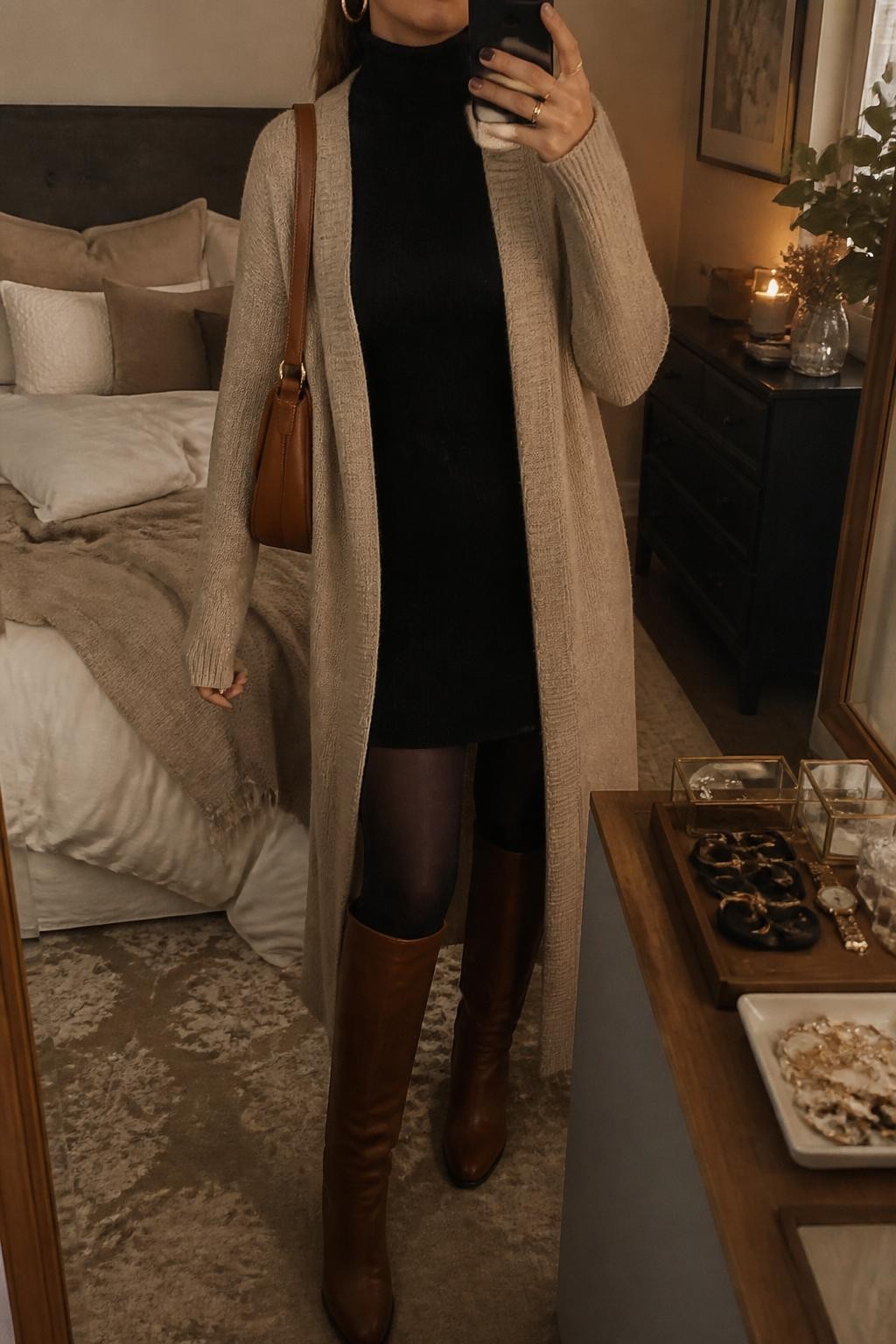 Mirror selfie of a woman wearing an oatmeal long maxi cardigan, black turtleneck sweater dress, black opaque tights, brown knee-high boots, gold hoop earrings, and a caramel shoulder bag, holding a phone in front of a mirror, cropped at NECK so the face does not show, relaxed casual selfie pose, in a cozy bedroom mirror setup, warm moody lighting, with a jewelry tray on the vanity and soft pillows on an unmade bed, latest iPhone photo quality.