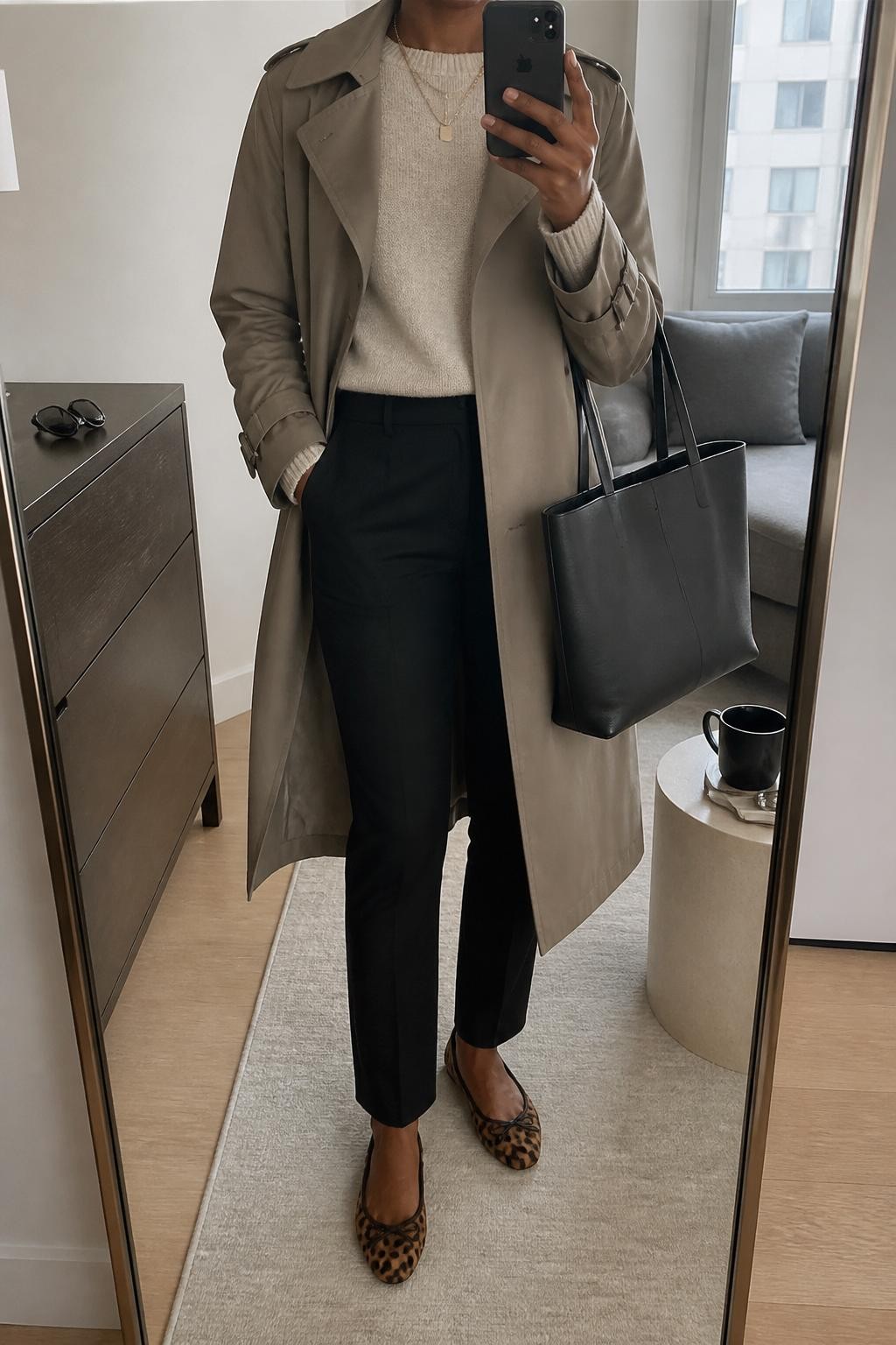 Mirror selfie of a Black woman wearing a taupe trench coat over a beige fine-knit sweater, black slim trousers, leopard print flats, gold layered necklace, and a black work tote, cropped at neck so the face does not show, in a stylish city apartment mirror with natural window light, sunglasses on a dresser and a coffee mug on a side table, realistic latest iPhone photo quality.