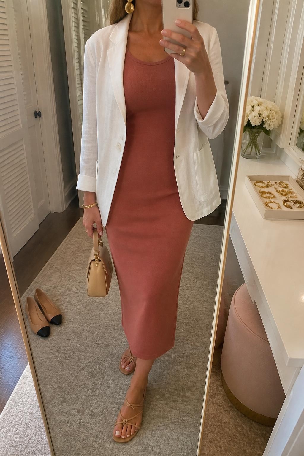 mirror selfie of a woman wearing the exact outfit described above, clearly showing a terracotta sleeveless column midi dress, white lightweight linen blazer worn open, nude strappy flat sandals, a tan mini handbag, and gold statement earrings, holding a phone in front of a mirror, cropped at NECK so the face does not show, with a relaxed casual selfie pose, in a feminine dressing room mirror, soft golden hour light, with a jewelry tray on the vanity and a pair of flats on the floor nearby, latest iPhone photo quality.